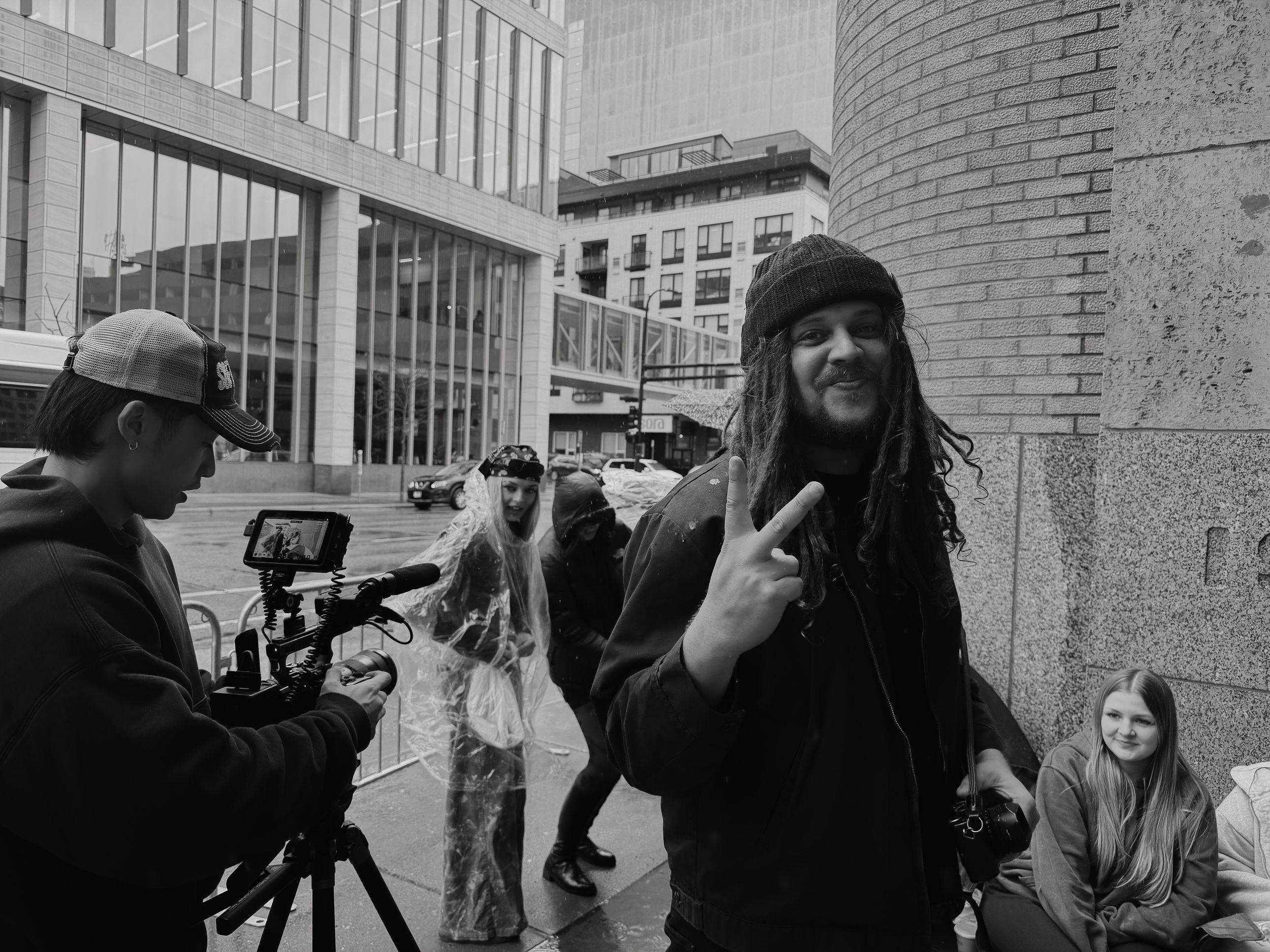 Black and white street photo of dreadlocked person gesturing at camera crew outdoors.