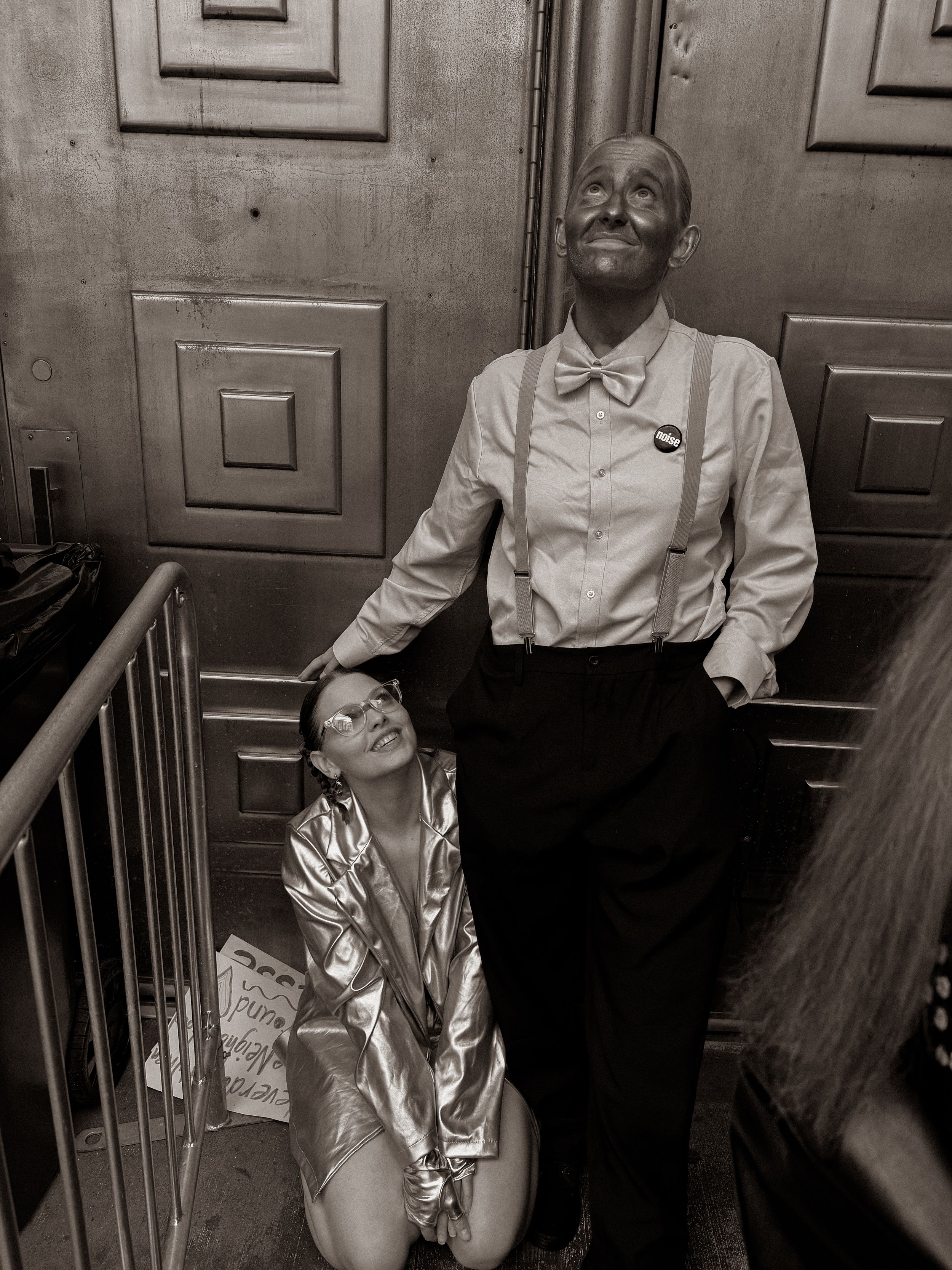 Black and white photo of two costumed performers on stairs, one kneeling in silver jacket, one standing in bow tie and suspenders.