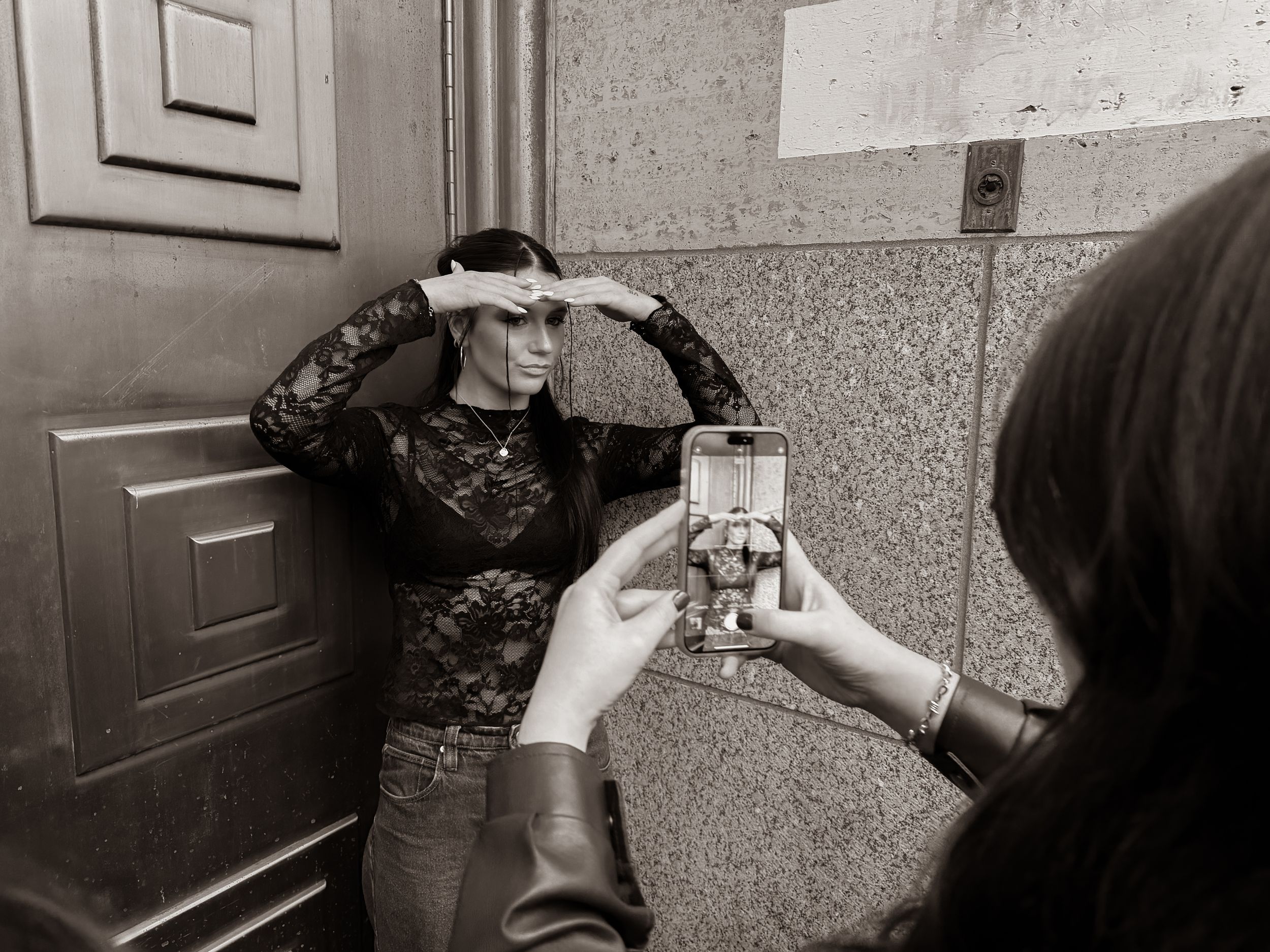 Black and white photo of a person posing against a wall while someone photographs them with a smartphone.