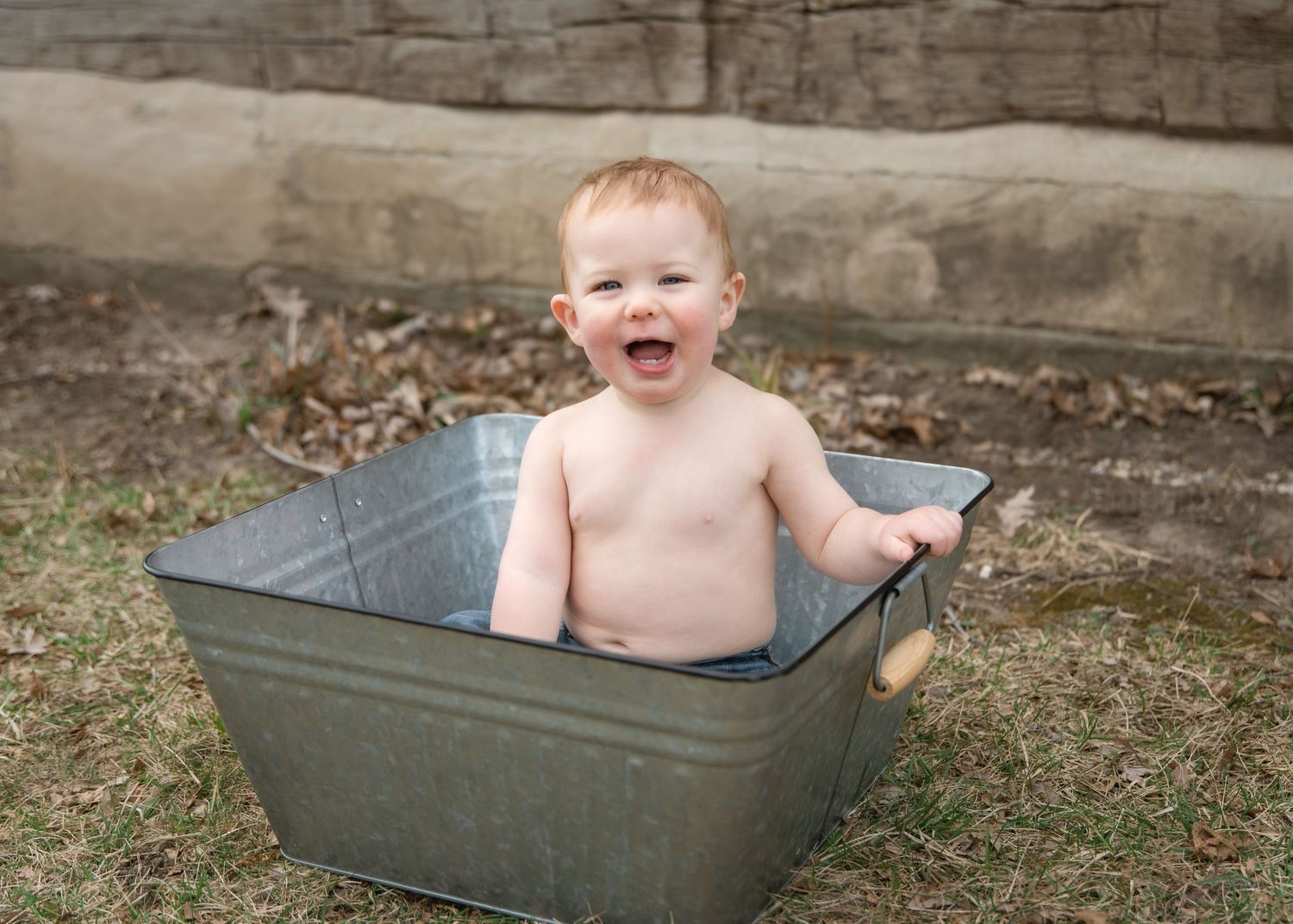 Happy baby sitting in a vintage galvanized metal tub outdoors, laughing joyfully in a sunny backyard setting.
