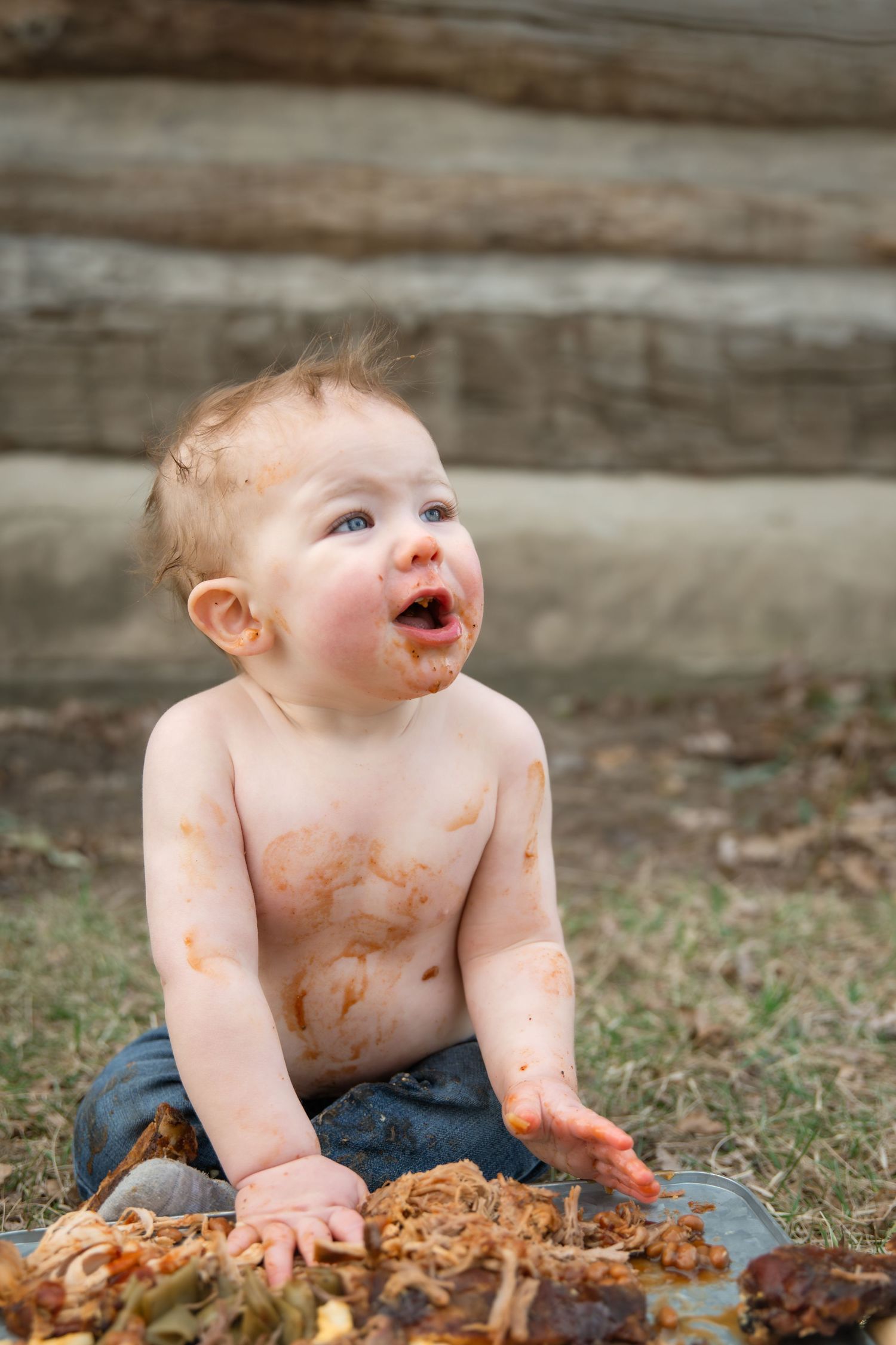 Messy baby covered in food sitting outdoors on grass, laughing with mouth open near wooden steps.