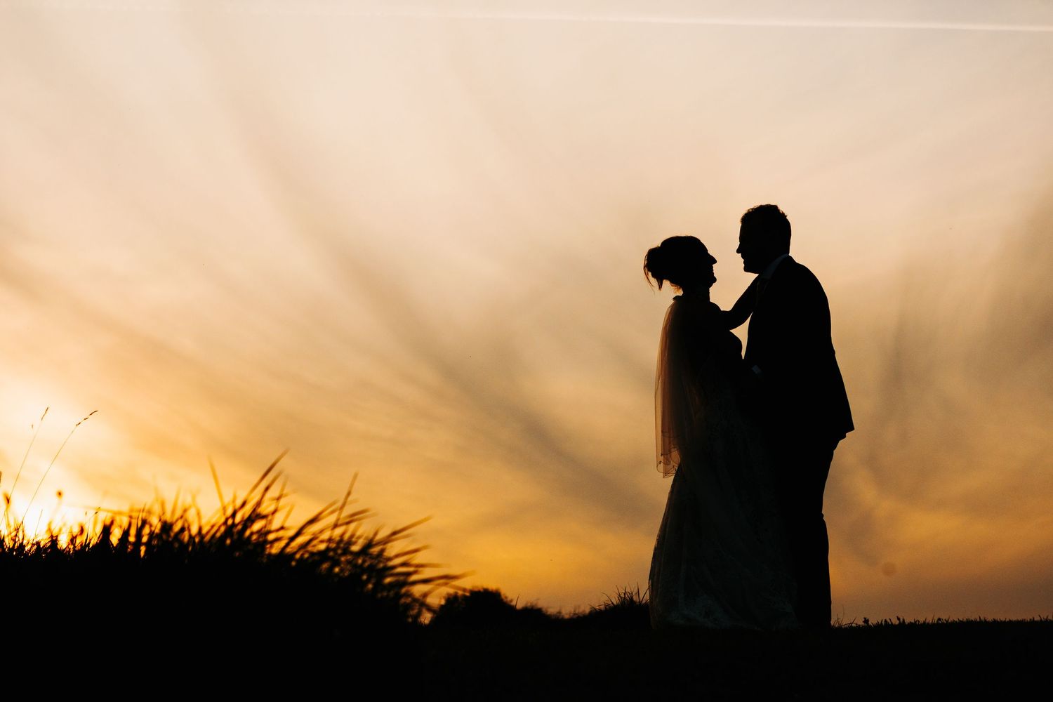 Silhouette of a bride and groom embracing at sunset with dramatic golden rays in the sky.