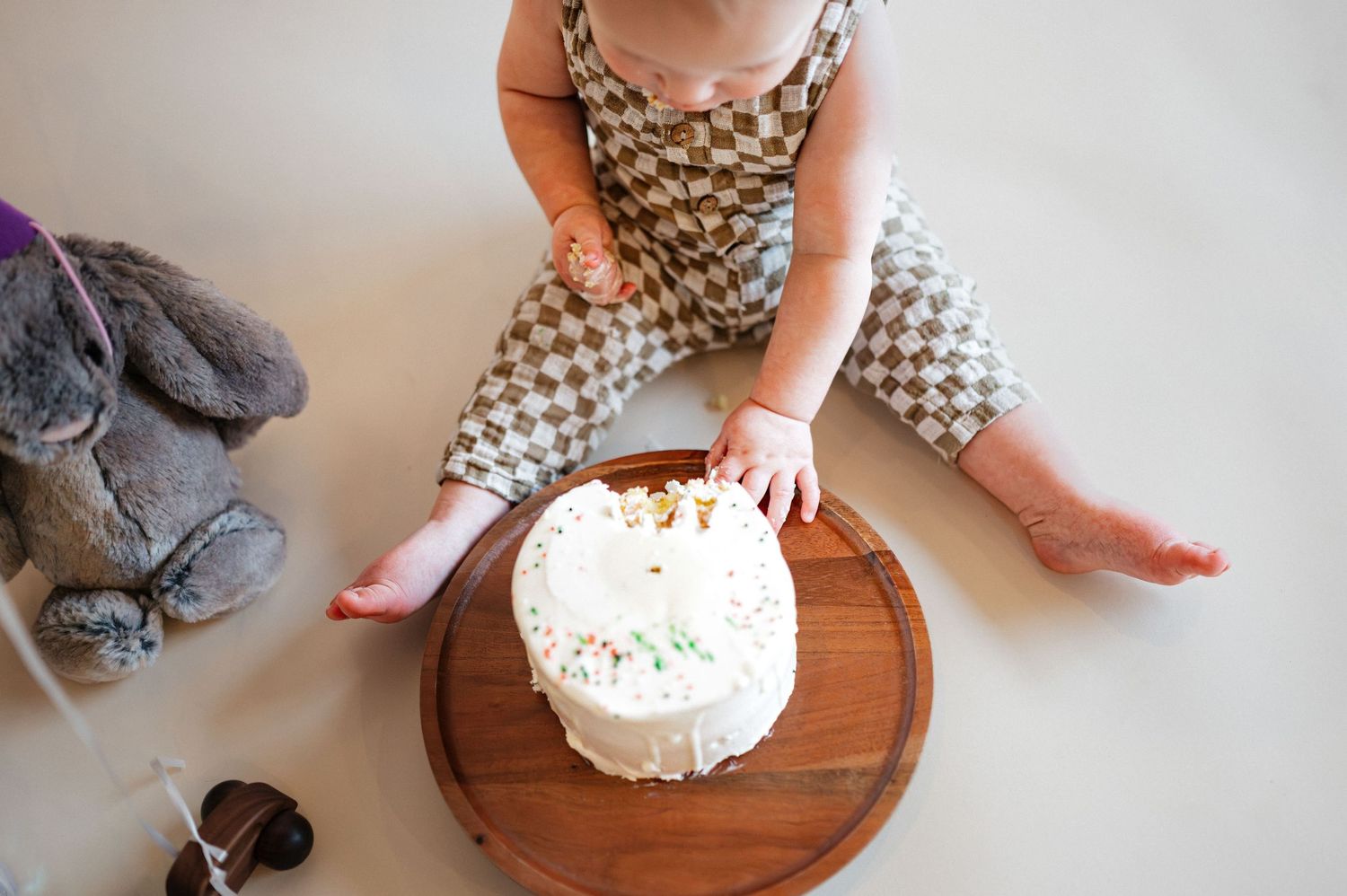 Baby in checkered romper touching a white smash cake on a wooden board during first birthday celebration.