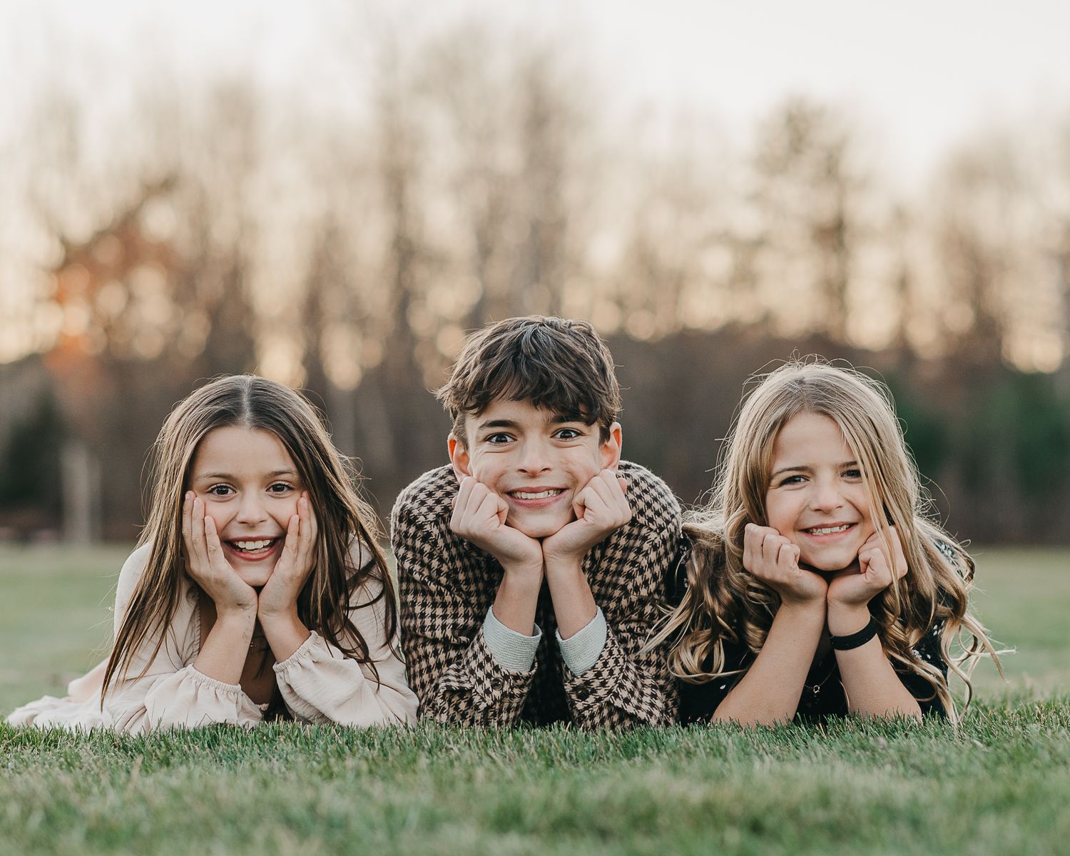 Three happy children lying on green grass in a park, smiling at camera during golden hour sunset.