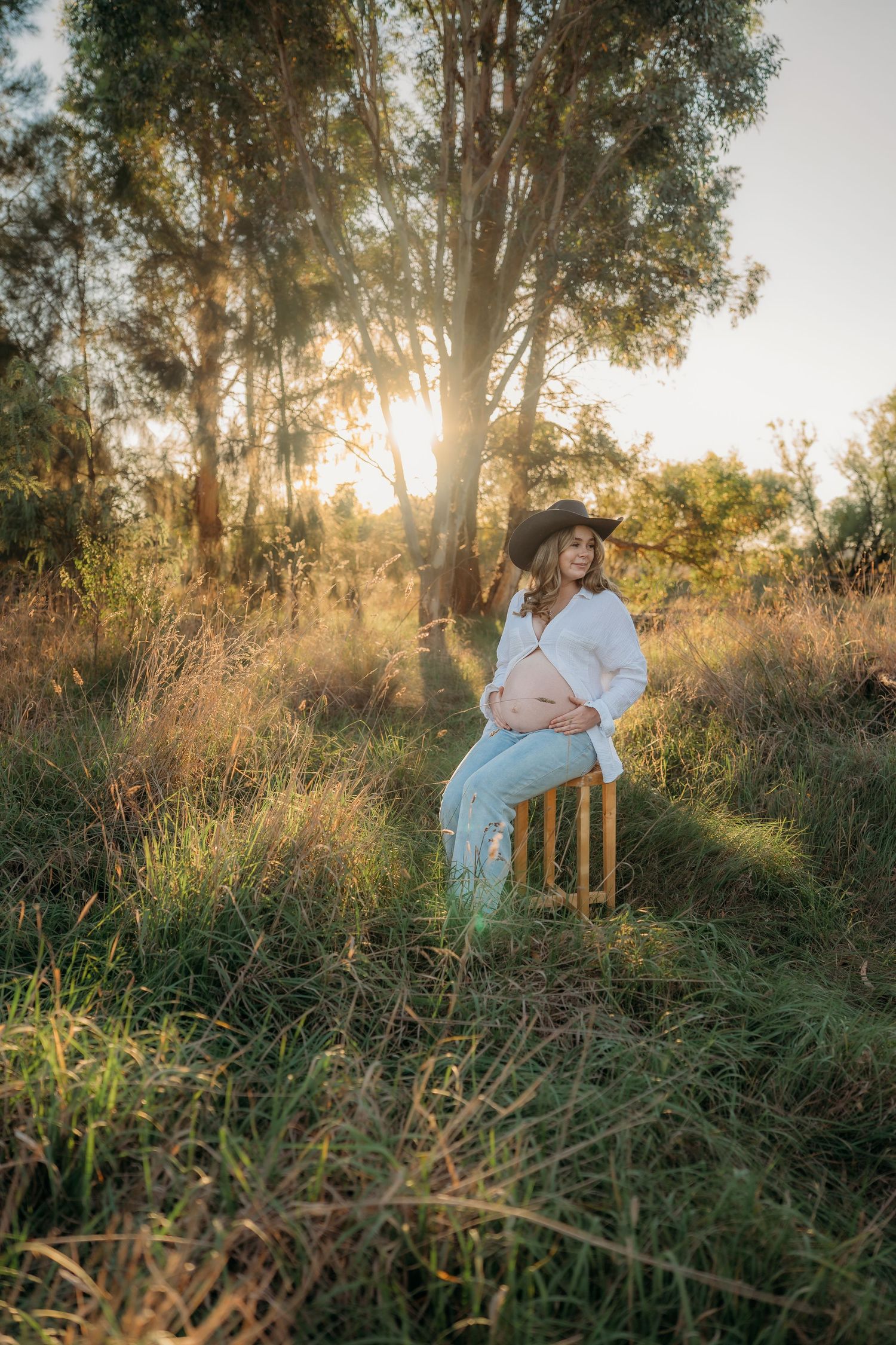 Pregnant woman in white shirt and cowboy hat sits on wooden chair in golden sunlit field surrounded by tall grass and trees.