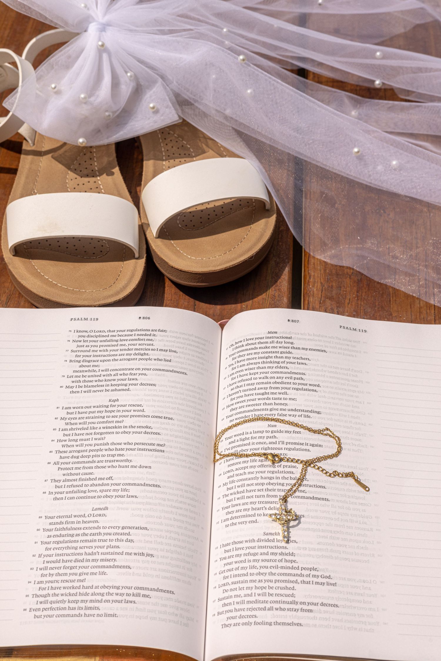 Open Bible with gold cross necklace, white bridal sandals, and pearl-trimmed veil on wooden surface.