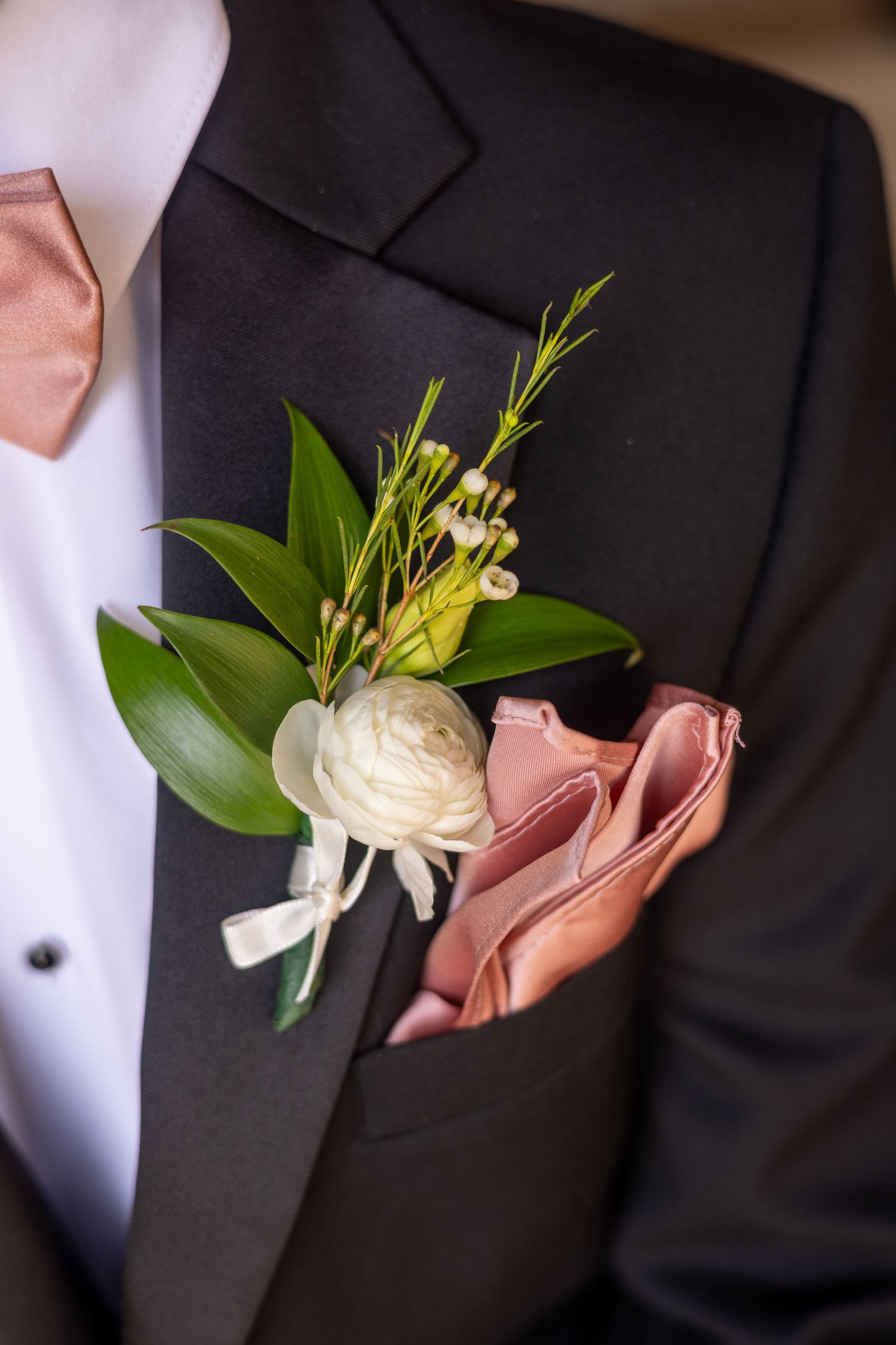 Close-up of a white ranunculus boutonniere with greenery on a black tuxedo with pink pocket square.