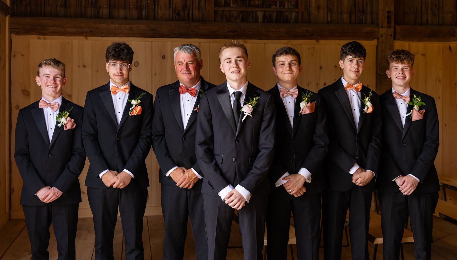Groom and groomsmen in black suits with floral boutonnieres posing inside a rustic wooden barn.