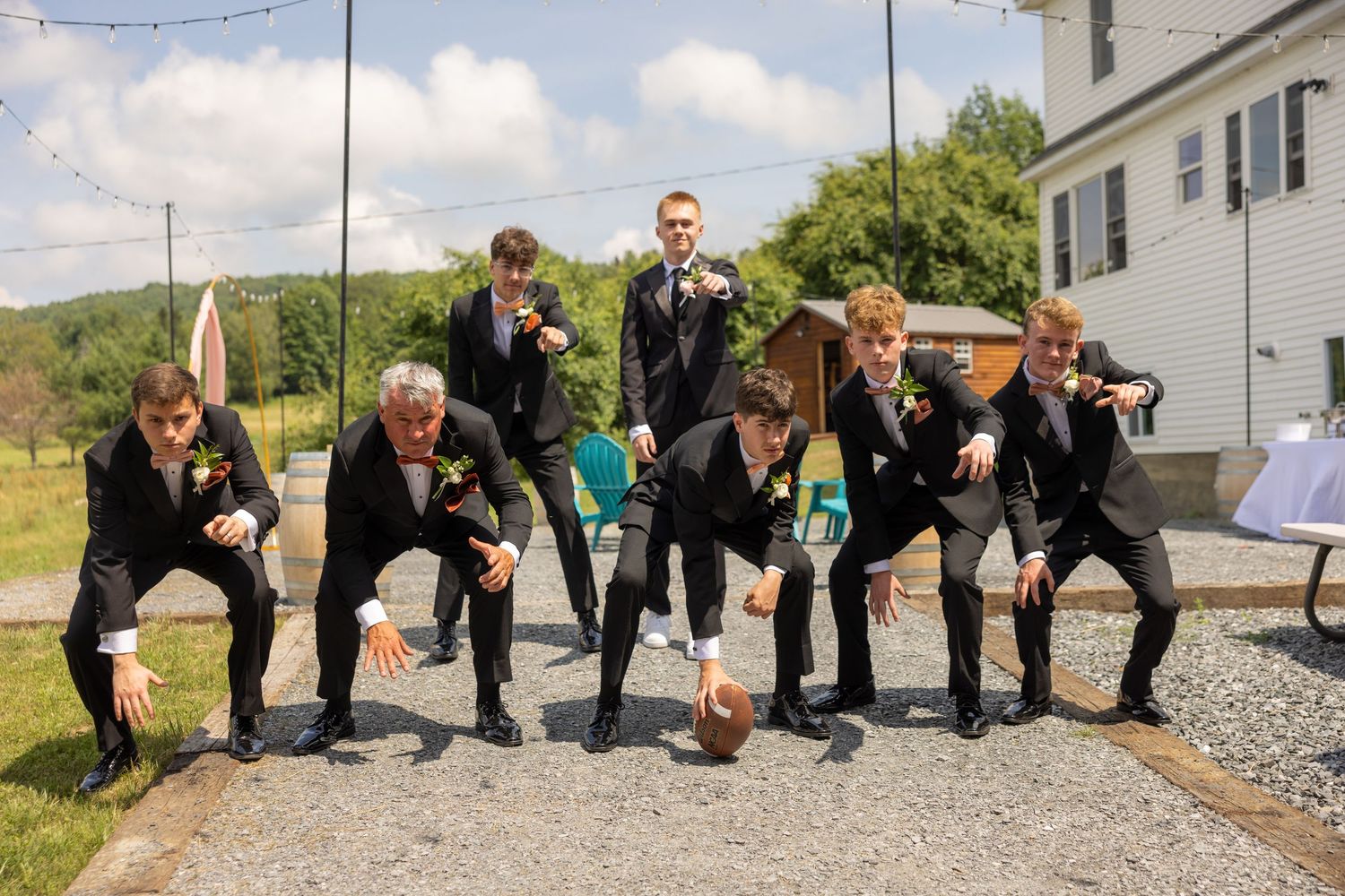 Groomsmen in black suits playfully pose with a football outside on a sunny wedding day.
