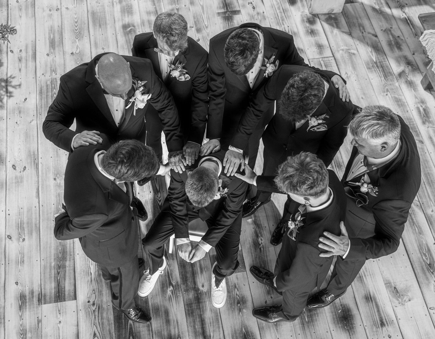 Aerial black and white photo of groomsmen gathered in a huddle on a wooden floor.