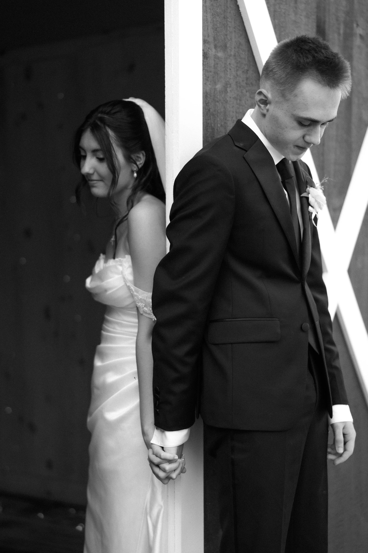 Black and white photo of bride and groom holding hands around a barn door pillar.