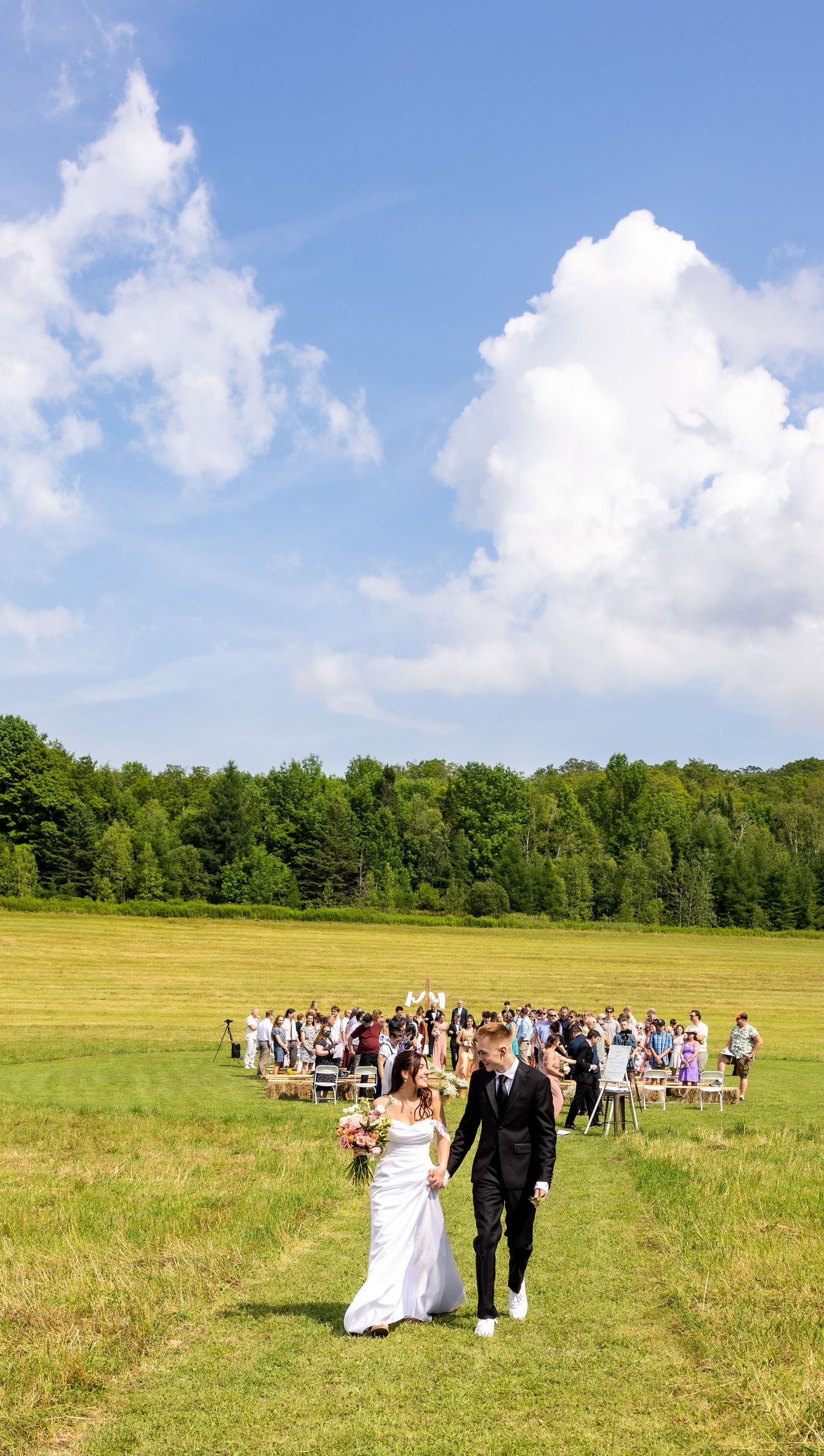 Newlyweds walk hand-in-hand through green field after outdoor wedding ceremony with guests and blue sky behind.