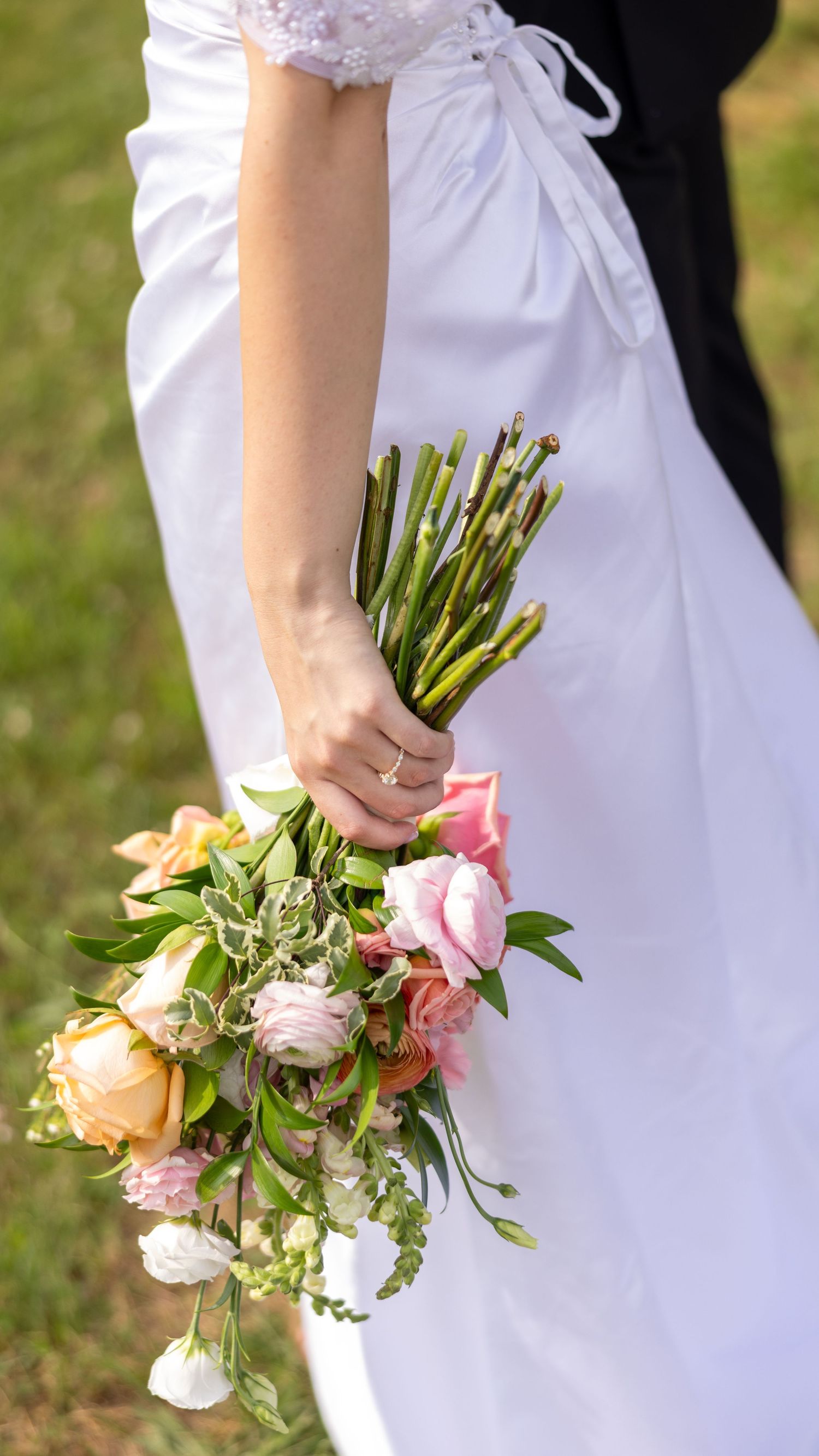 Bride in white wedding gown holds pink and peach rose bouquet at her side on green lawn.