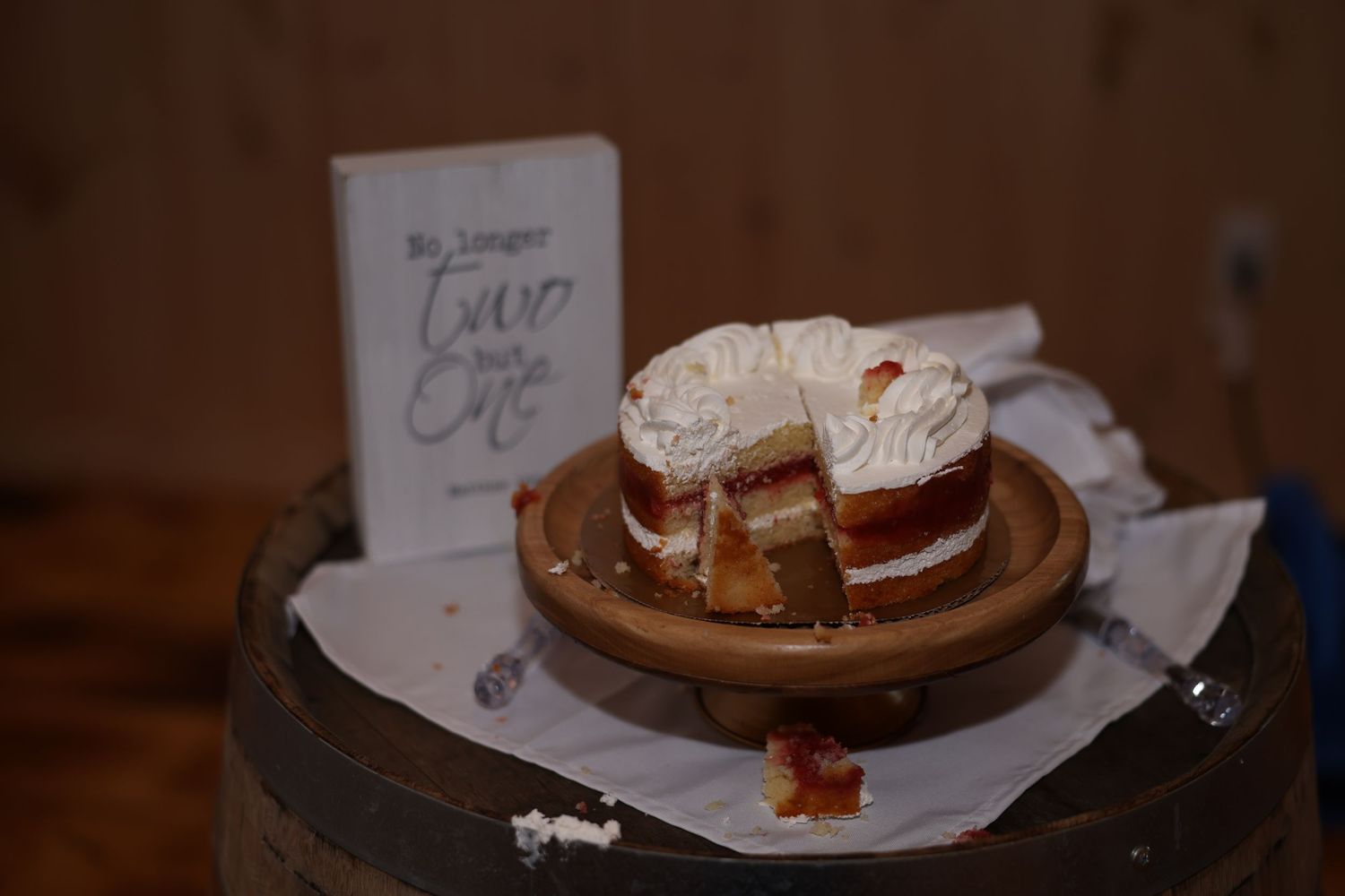 Partially sliced rustic naked wedding cake on wooden barrel with decorative sign at reception venue.