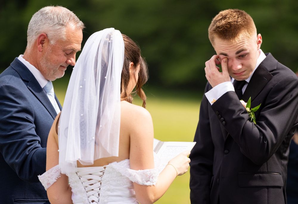 Emotional groom wiping tears during outdoor wedding ceremony with officiant and bride.