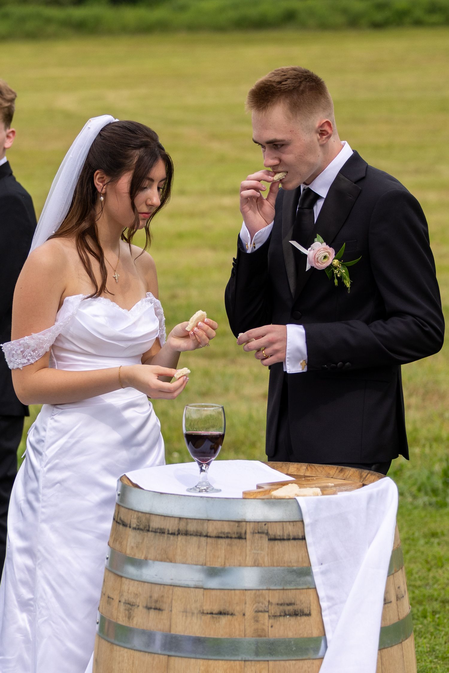 Bride and groom sharing food at outdoor wedding ceremony beside a wine barrel with red wine.