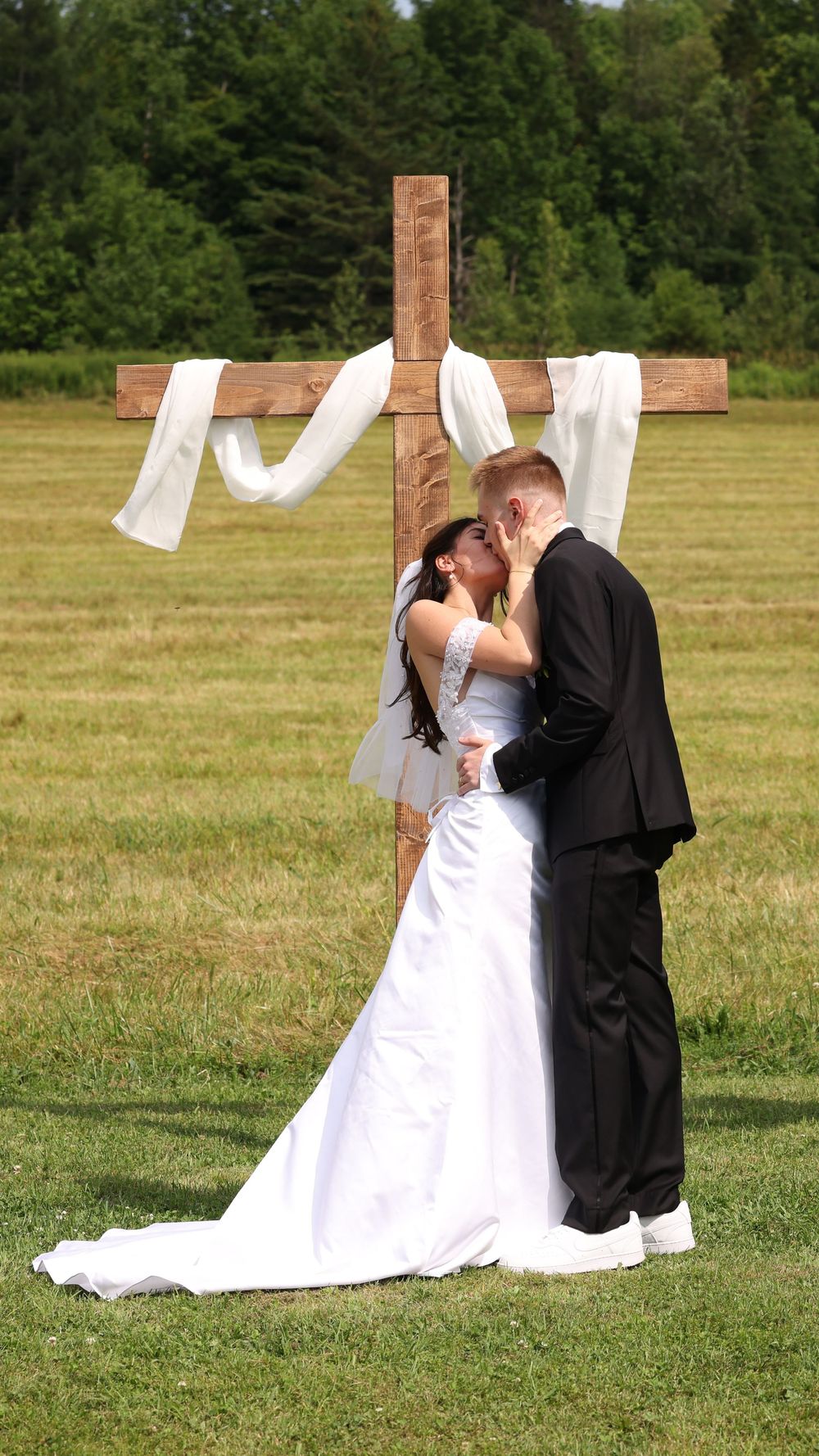 Bride and groom sharing first kiss in front of wooden cross at outdoor wedding.