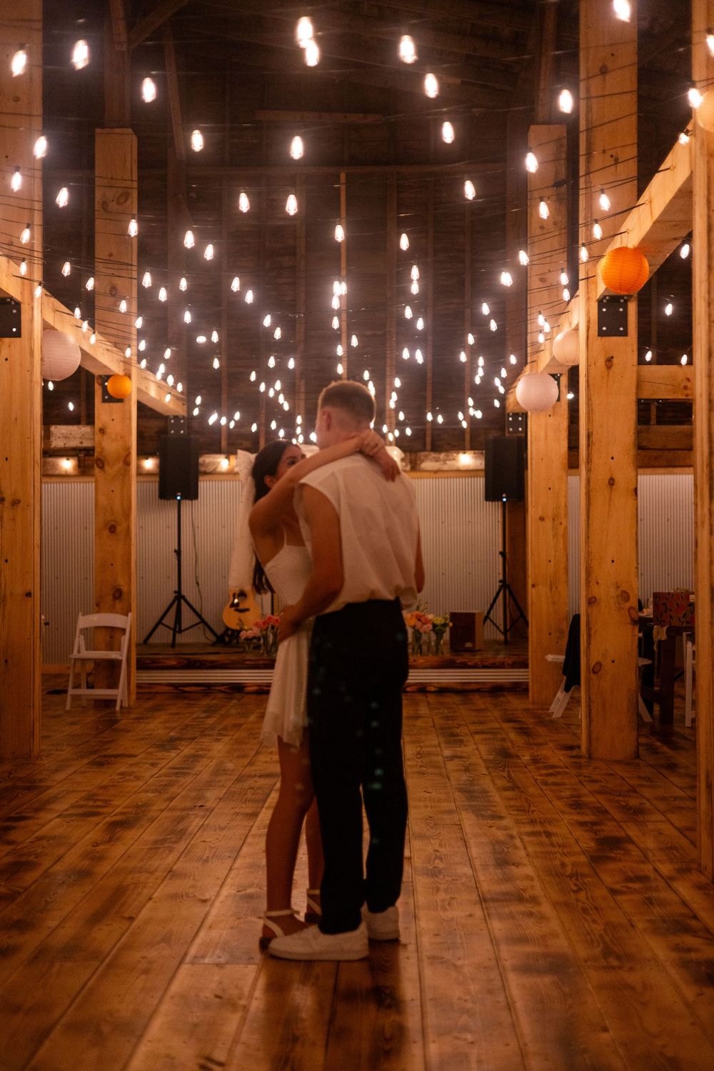 Newlyweds sharing first dance alone in rustic barn adorned with string lights.