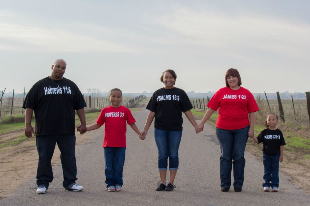 A family of five wearing black and red shirts with scripture, holding hands while standing in a line.