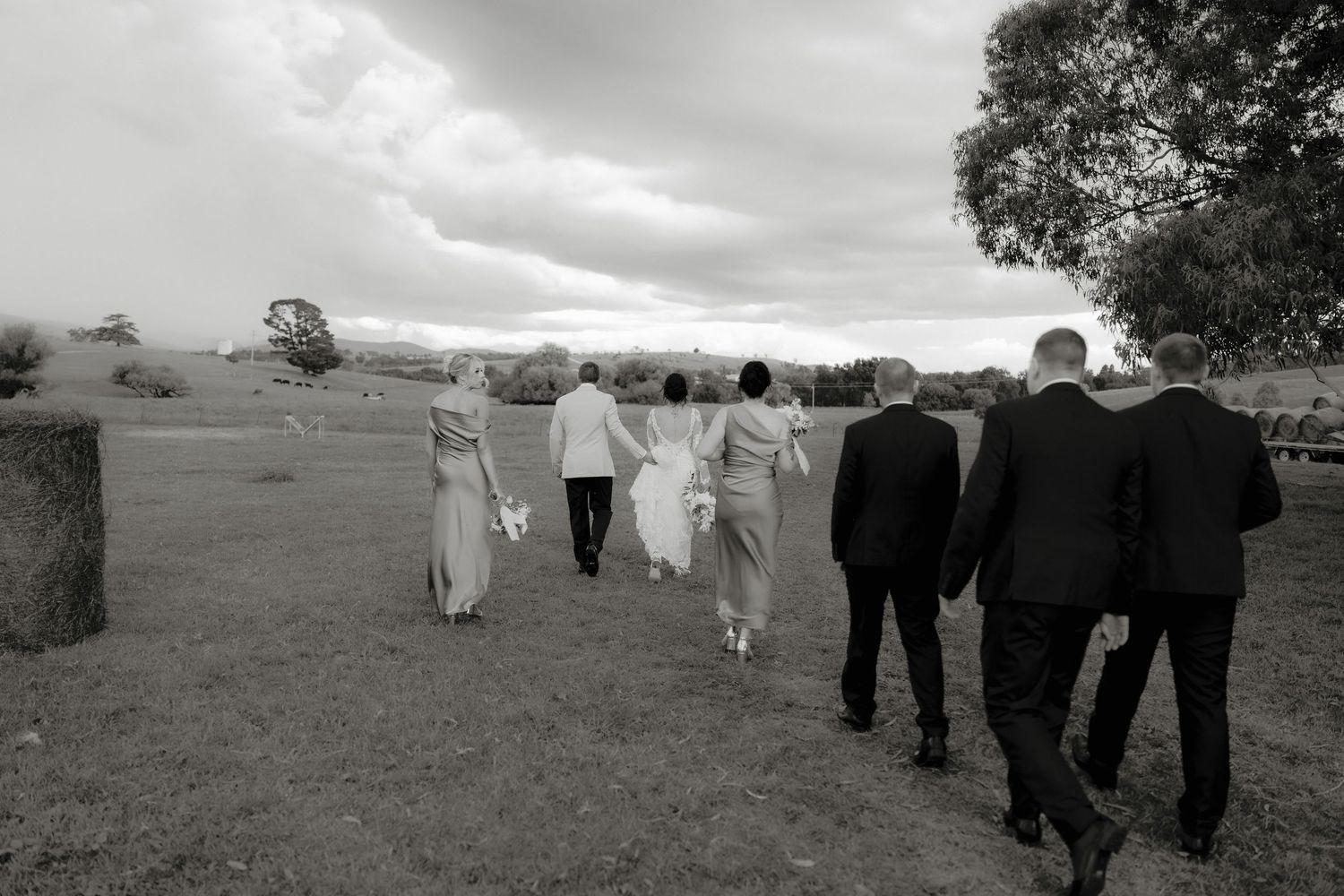 Black and white photo of a wedding party walking across a rural field with hay bales and dramatic cloudy sky.
