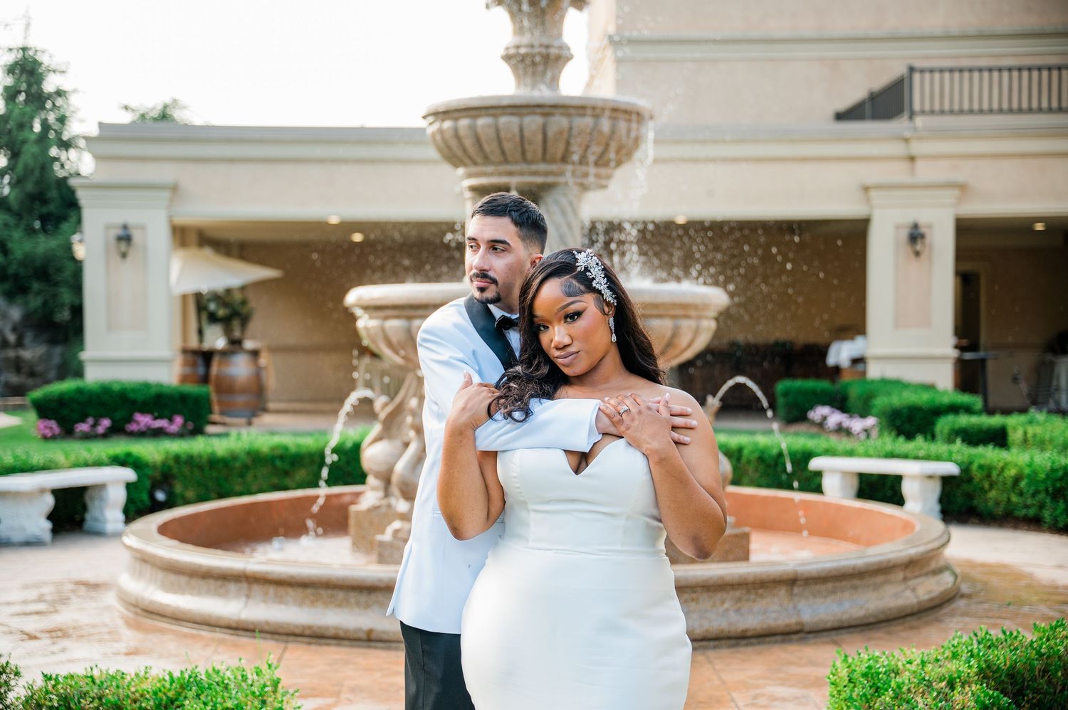 Couple embracing in front of elegant fountain at wedding venue, bride in white gown, groom in formal attire at Aria in Prospect, CT.