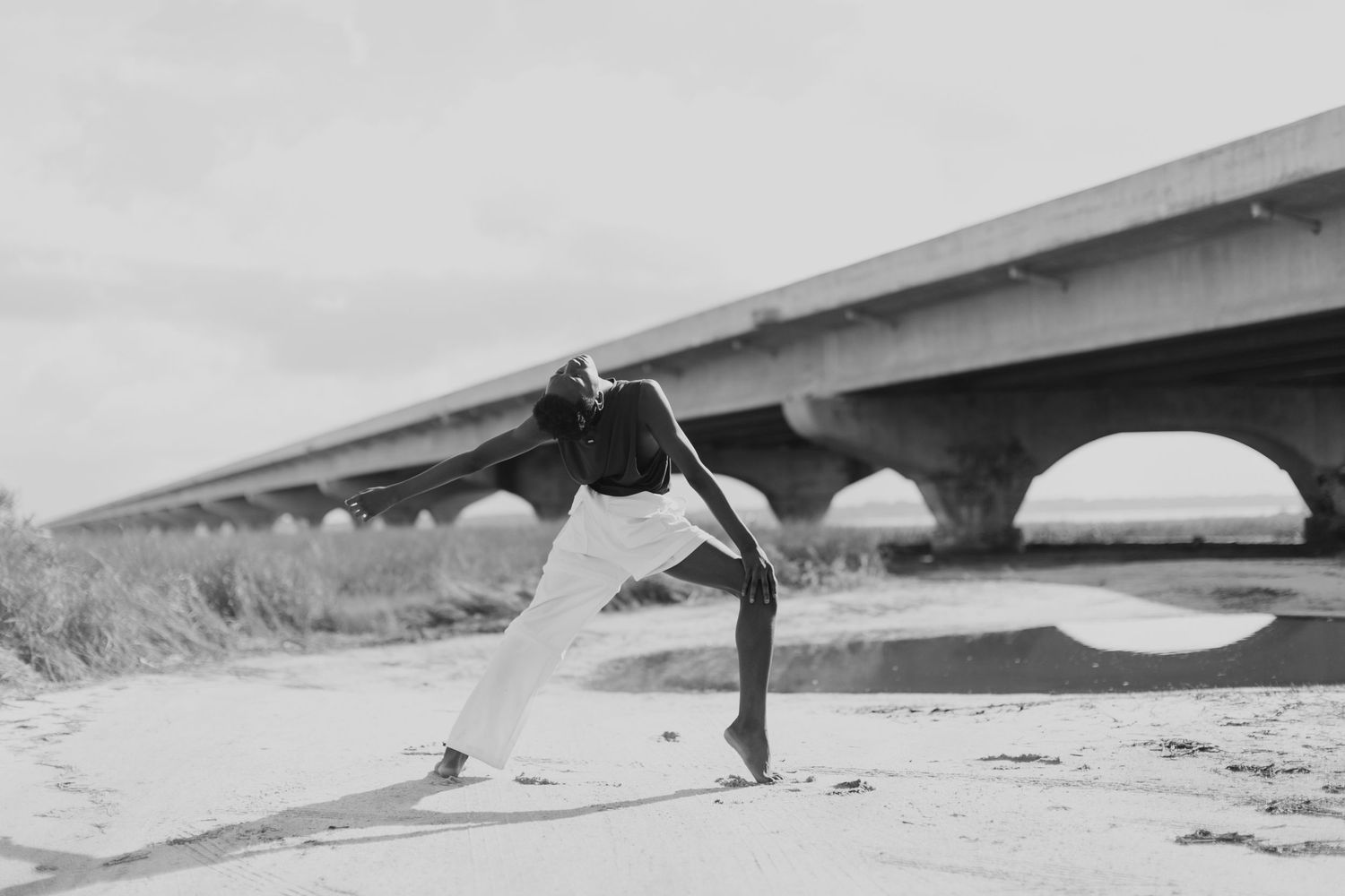 Black and white photo of a dancer gracefully balancing under a concrete bridge on sandy ground.