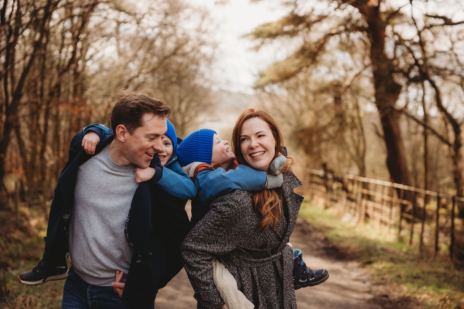 Family of four enjoying countryside views by a rustic gate, with father pointing at scenic landscape on a sunny winter day.