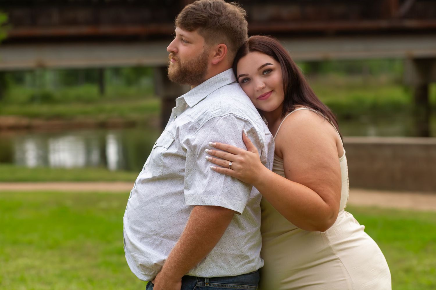 Couple posing outdoors near a bridge, woman in cream dress hugging man in white shirt from behind.