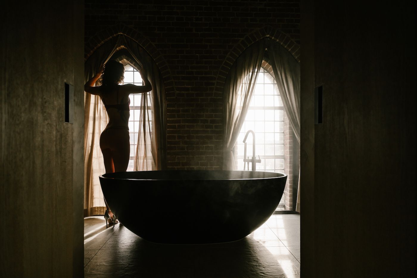Silhouette of a woman standing by arched windows near a freestanding black bathtub in a moody bathroom.