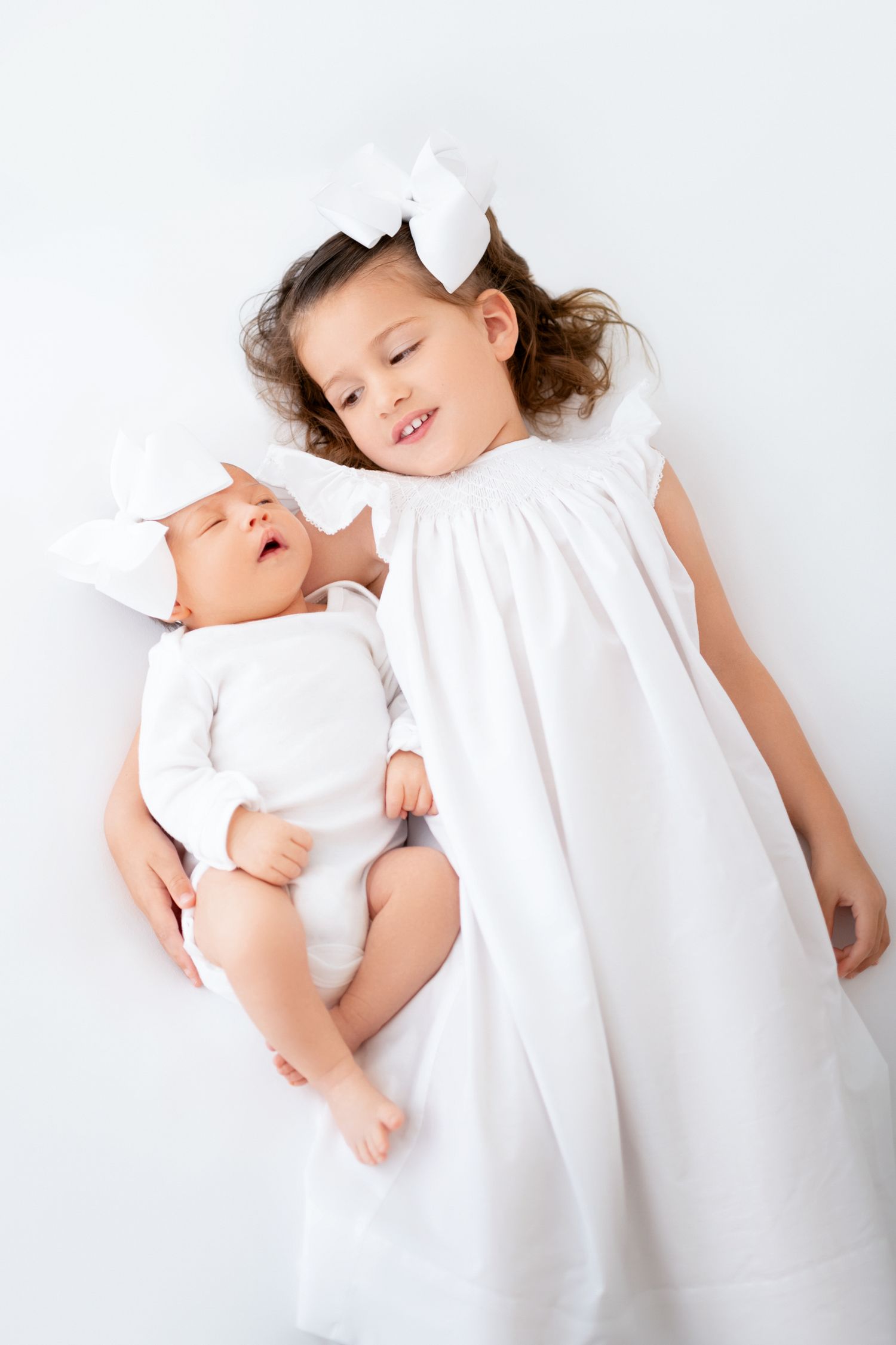 Older sister cuddling newborn baby girl during a soft white studio newborn photography session in Winchester, Virginia
