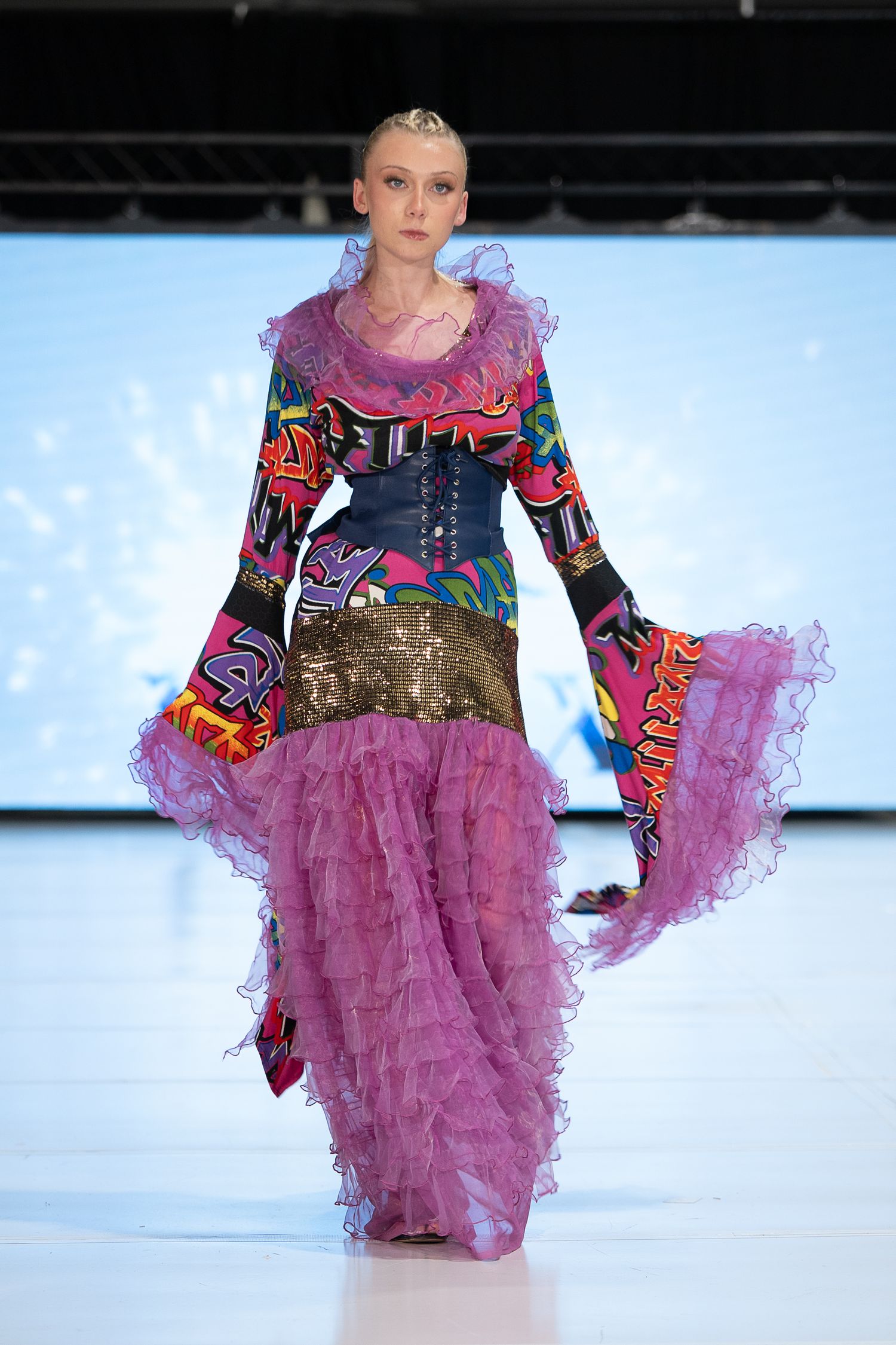 Model walks runway in vibrant multicolor printed gown with pink ruffled skirt and gold sequin waistband at fashion show.