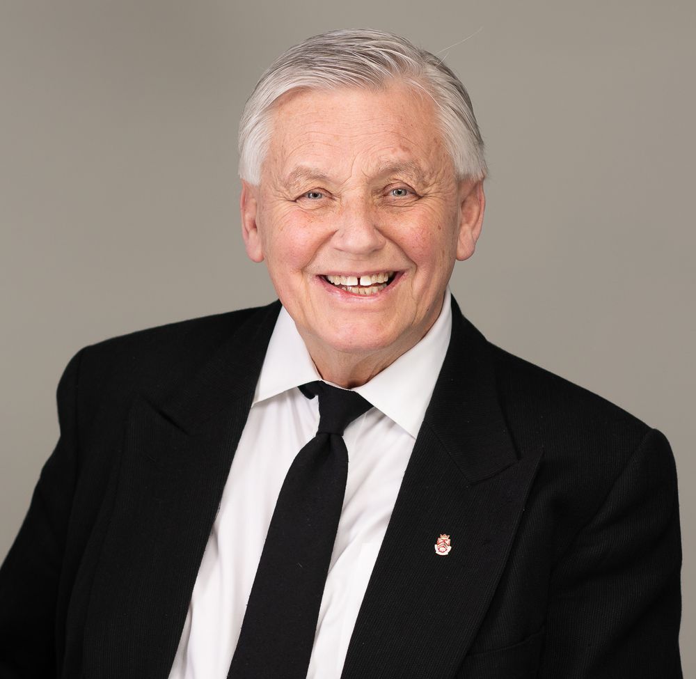 Smiling senior man with white hair wearing a black suit and tie with a lapel pin, professional headshot on gray background.