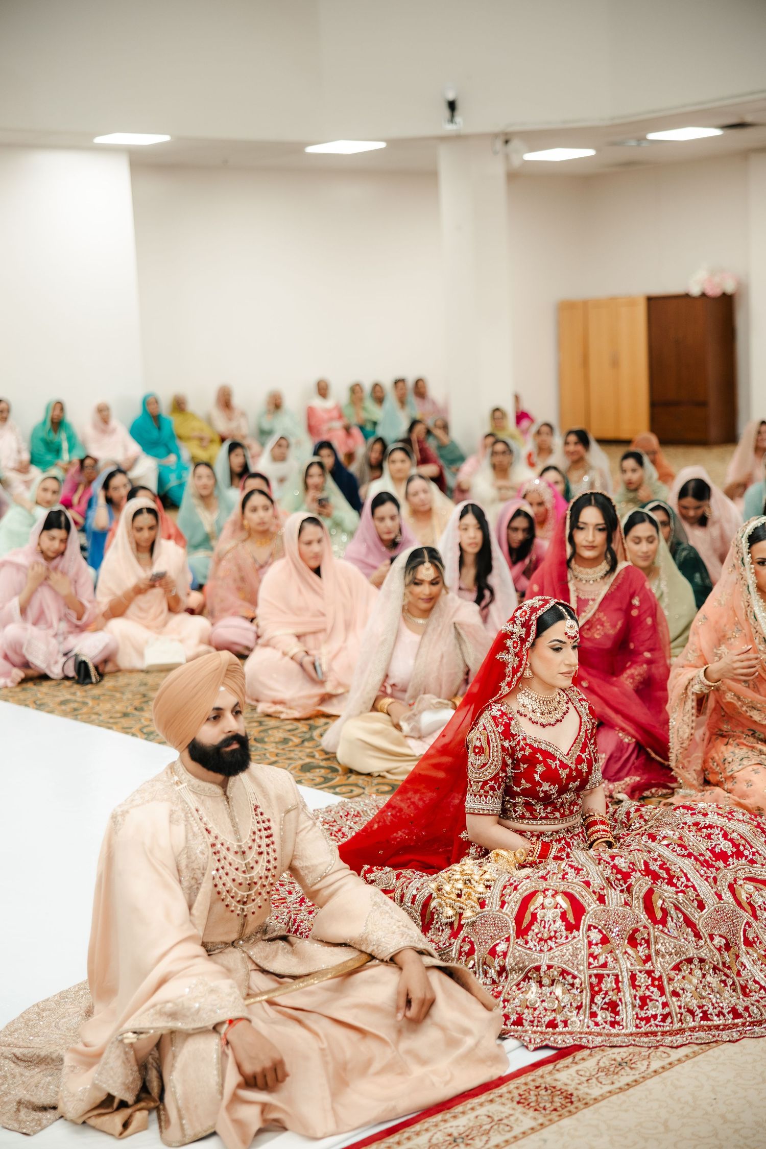 Sikh wedding ceremony with bride in red lehenga and groom in beige sherwani seated before congregation in gurudwara.