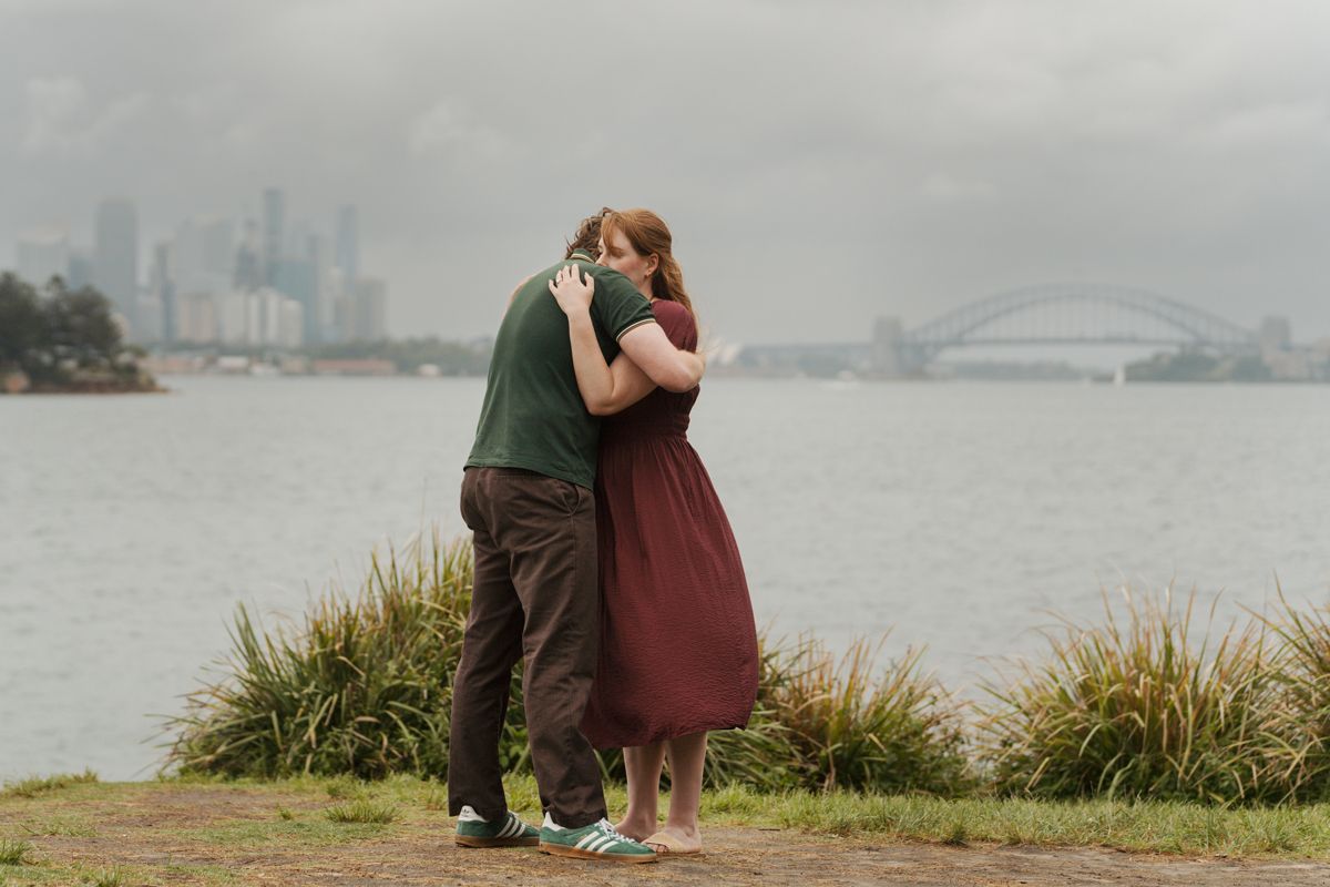 Couple hugging near Sydney Harbour with the iconic bridge and city skyline visible on a cloudy day during an engagement photoshoot