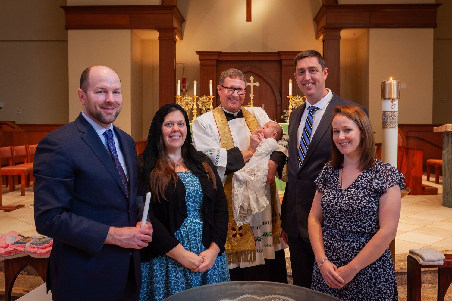 Group of five adults pose with a priest holding a baby in white baptismal gown at church.