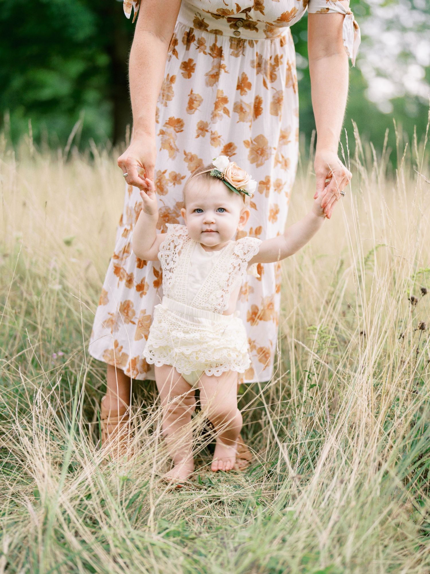 A smiling baby in a lace romper stands in a golden field, held by a woman in a floral dress.