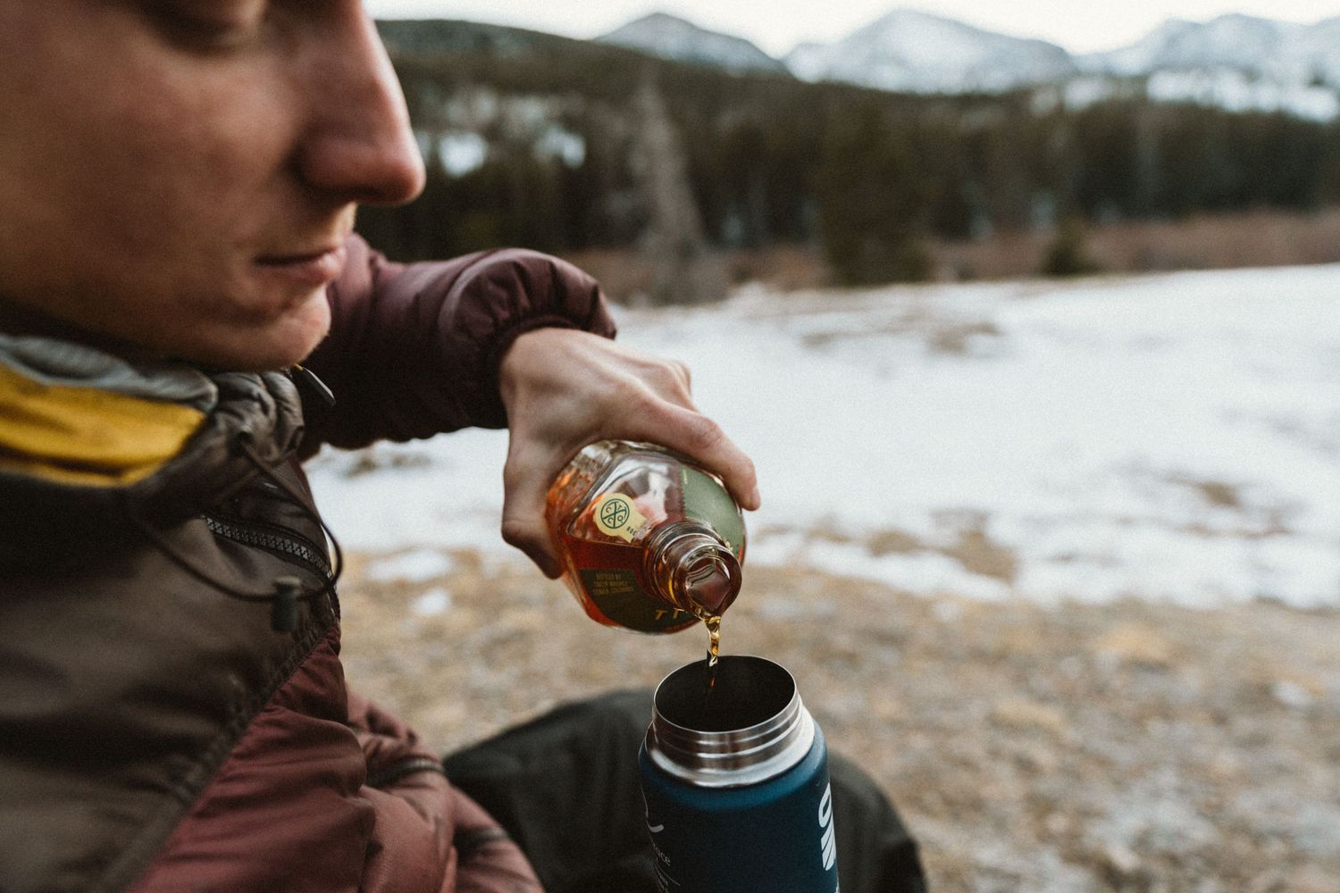 Person pouring hot beverage from thermos in snowy mountain setting.