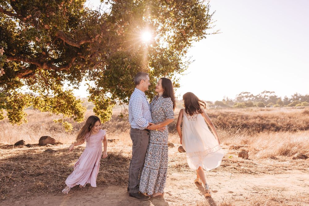 Family of four sharing a golden sunset moment under a tree in a sunlit field, children in pastel dresses.