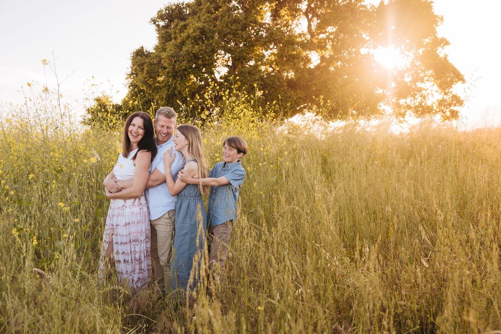 Family of four embracing in golden sunlit field with tall grass and trees during sunset outdoor photo session.