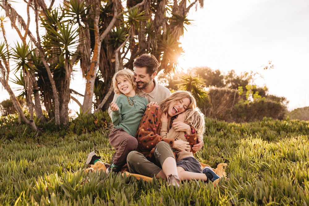 A happy family of four sits together on lush green grass, bathed in golden sunset light beneath tropical trees.