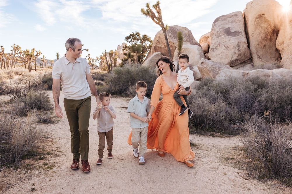 Family walking in Joshua Tree desert landscape, parents with two young boys, woman in orange maxi dress among rocky terrain.