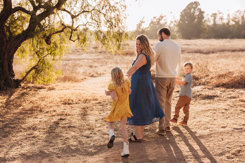Family of four walking on a sunlit dirt path surrounded by golden fields and trees during a warm sunset outdoor photoshoot.