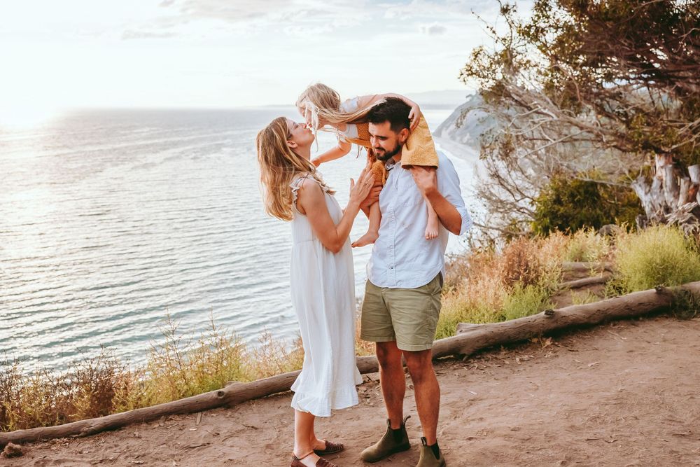 A happy family with a toddler on dad's shoulders enjoys a scenic coastal overlook at sunset.