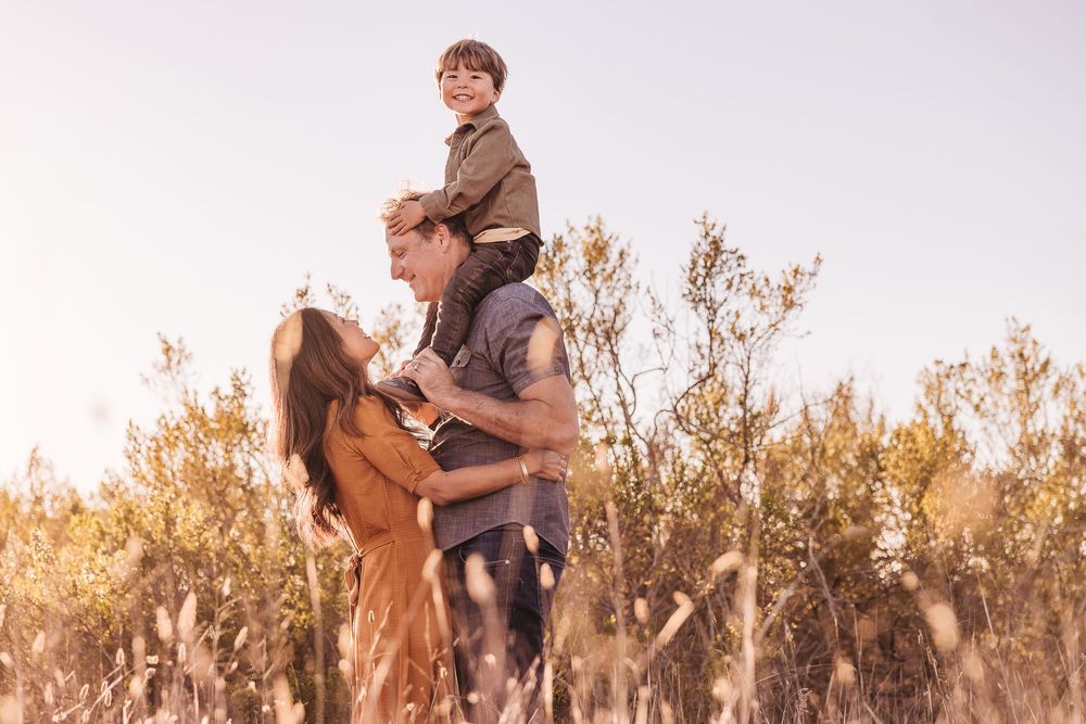 Family enjoying a sunny day outdoors, with a child sitting on a man's shoulders while a woman smiles beside them in a field.