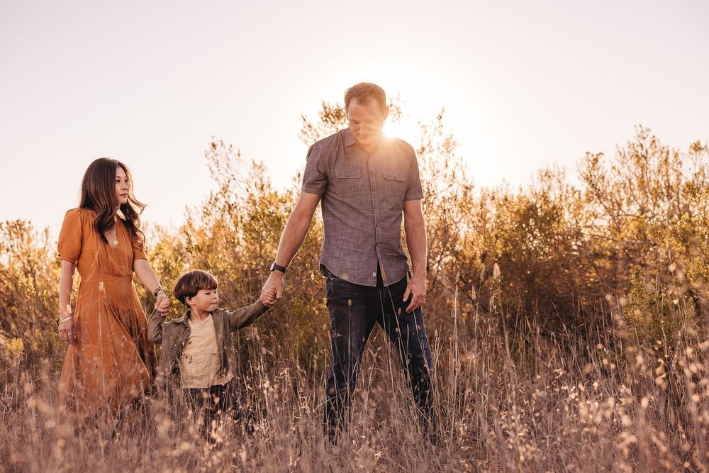 Family of three holding hands in a sunlit golden field, parents and toddler enjoying a warm sunset outdoors.