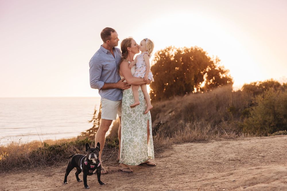 Family with toddler and black dog enjoying golden sunset on coastal bluff overlooking the ocean.