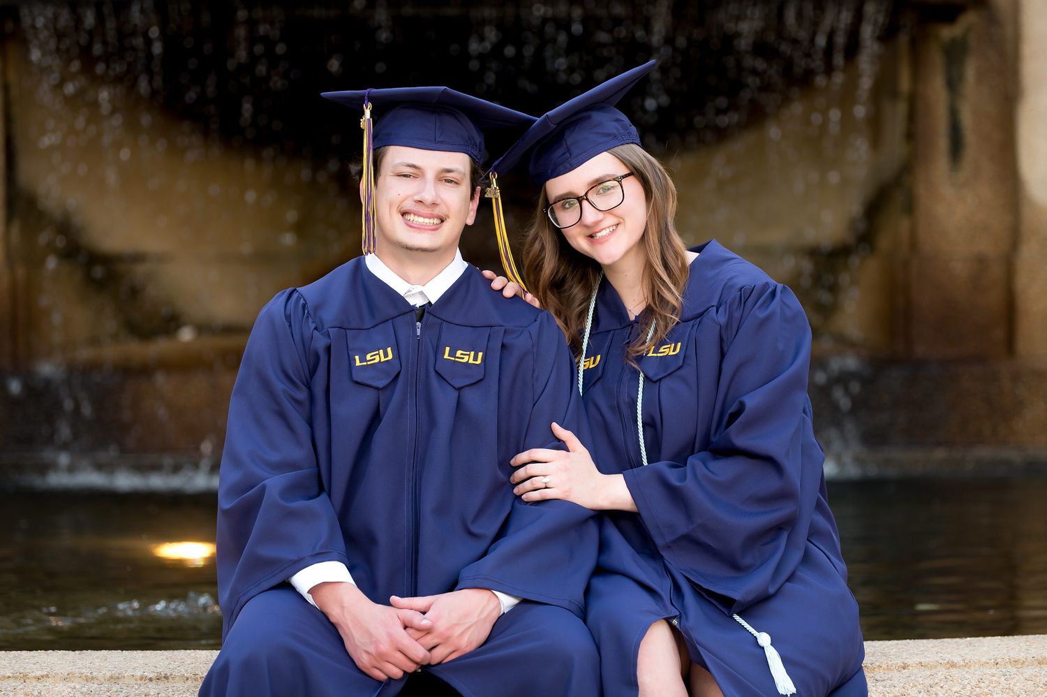 Two LSU graduates in navy caps and gowns smile together in front of a fountain.