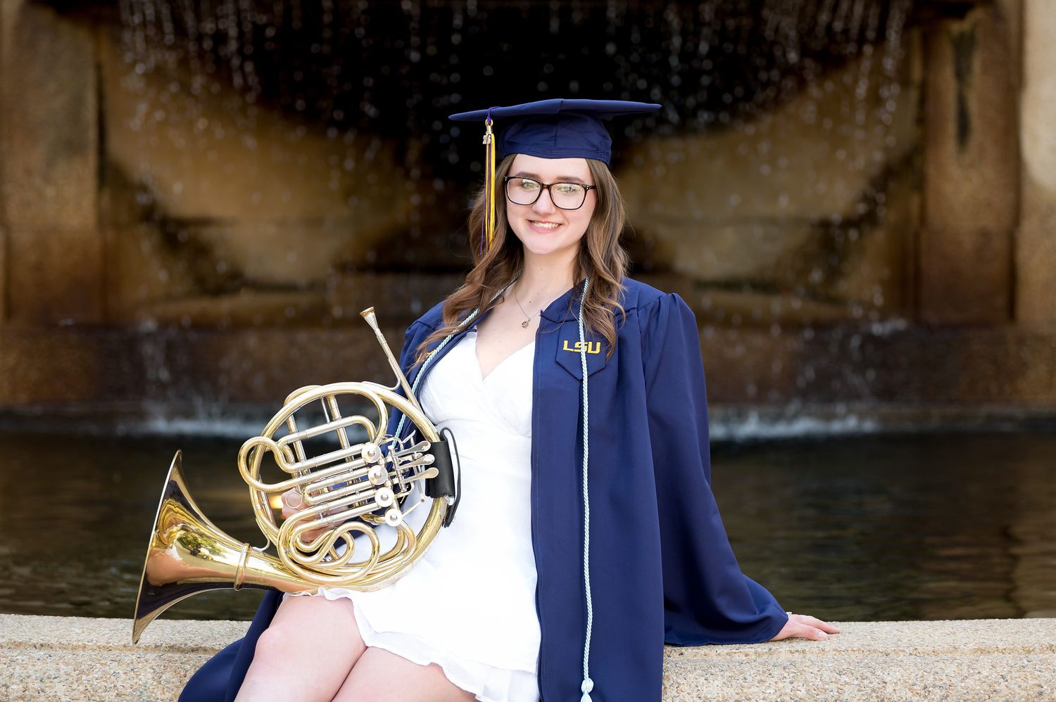 LSU graduate in navy cap and gown holds a French horn while sitting by a fountain.