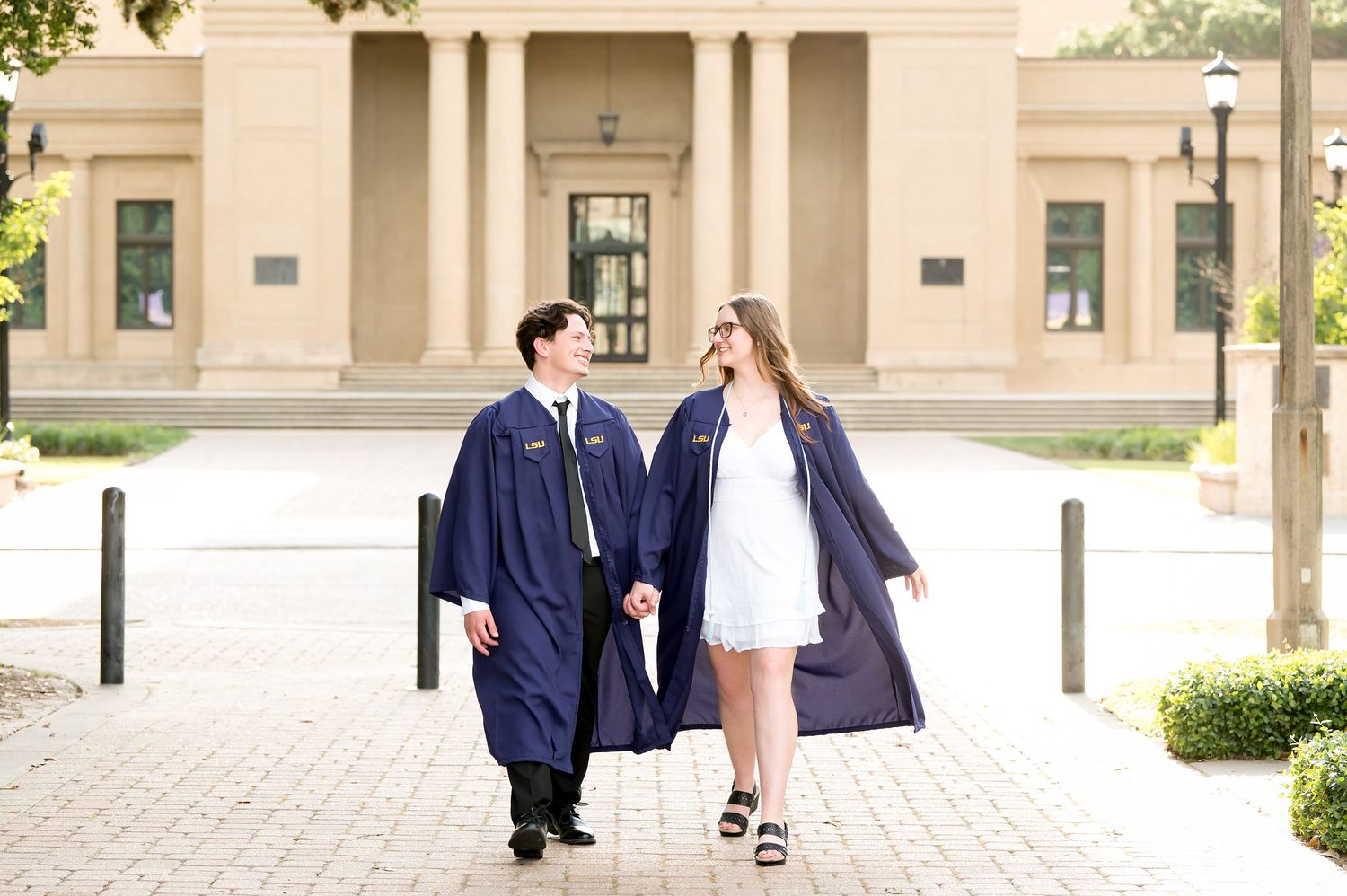 Two graduates in navy blue caps and gowns walk together smiling outside a classical building on graduation day.
