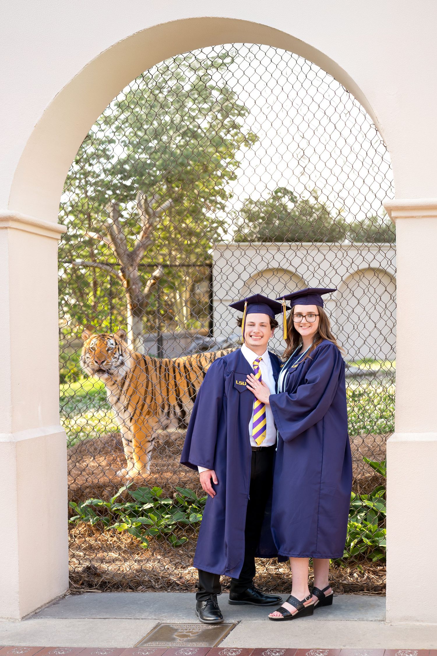 Two graduates in blue caps and gowns pose under an arch with a tiger visible behind a fence.